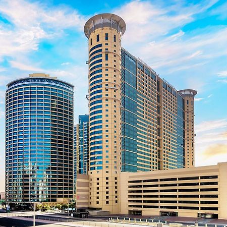 Grand Millennium Al Wahda Hotel And Executive Apartments Abu Dhabi Exterior photo The photo depicts a modern architectural landscape featuring two prominent high-rise buildings and a parking structure. The buildings exhibit a mix of glass and stone facades, showcasing contemporary design elements. The structure on the left is roun