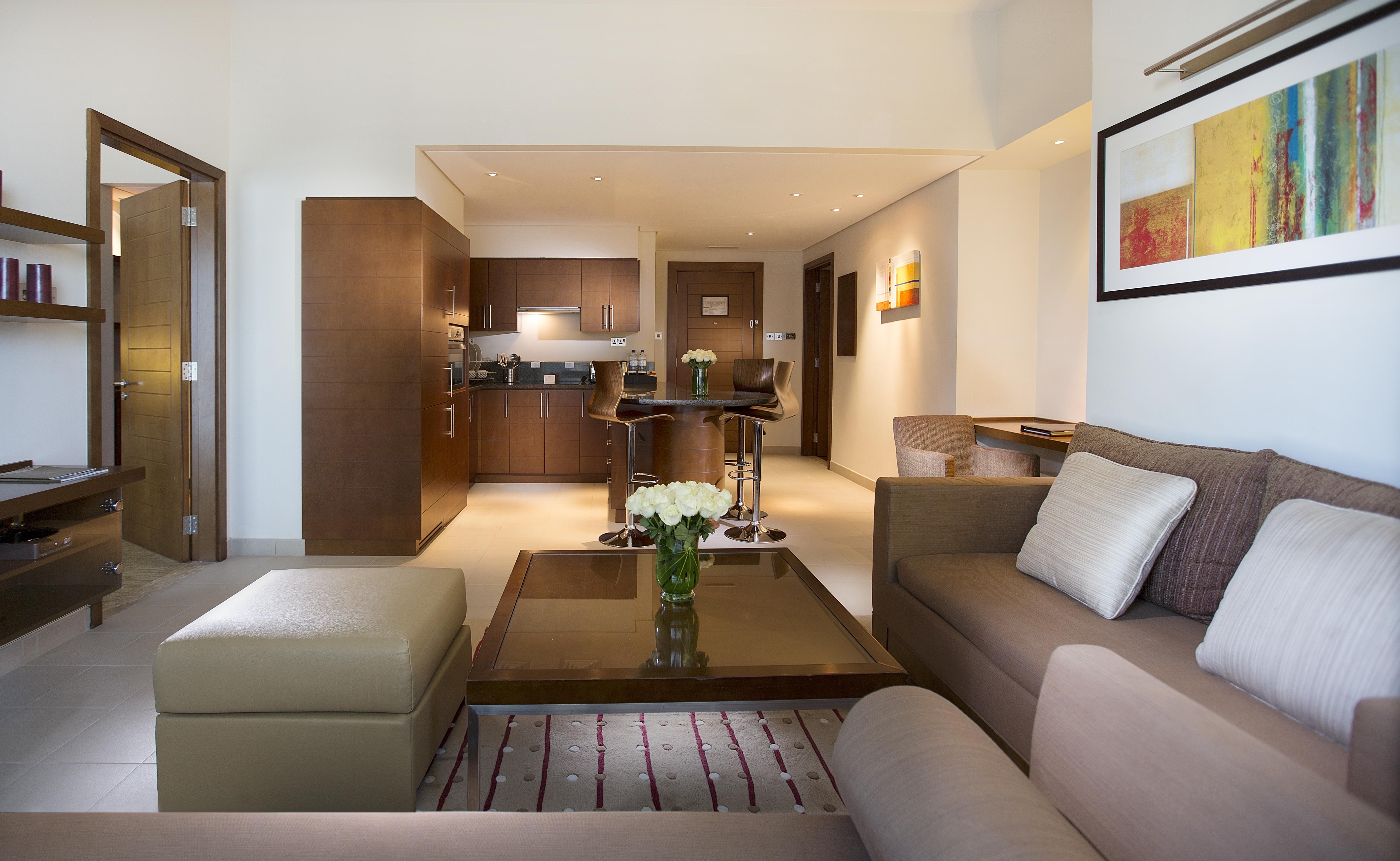 Grand Millennium Al Wahda Hotel And Executive Apartments Abu Dhabi Exterior photo The photo shows a modern living space that includes a comfortable sitting area and a kitchen. The sitting area features a soft beige sofa and an ottoman, with a coffee table in the center displaying a vase of flowers. The area is well-lit with natura