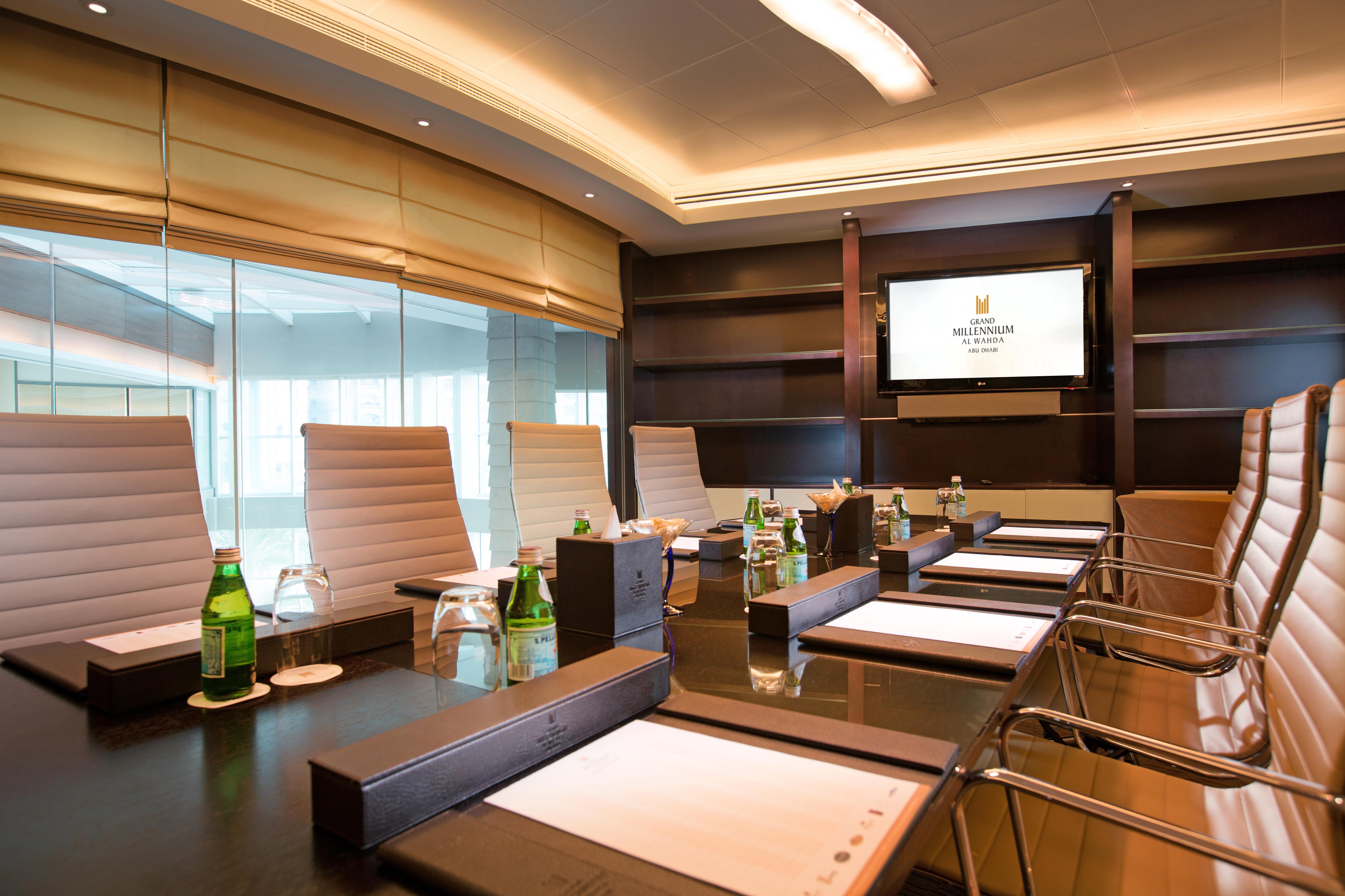 Grand Millennium Al Wahda Hotel And Executive Apartments Abu Dhabi Exterior photo The photo displays a modern conference room featuring a long, dark wooden table with several ergonomic chairs arranged around it. There are notepads and pens placed in front of each chair. In the background, there is a large screen mounted on the wal