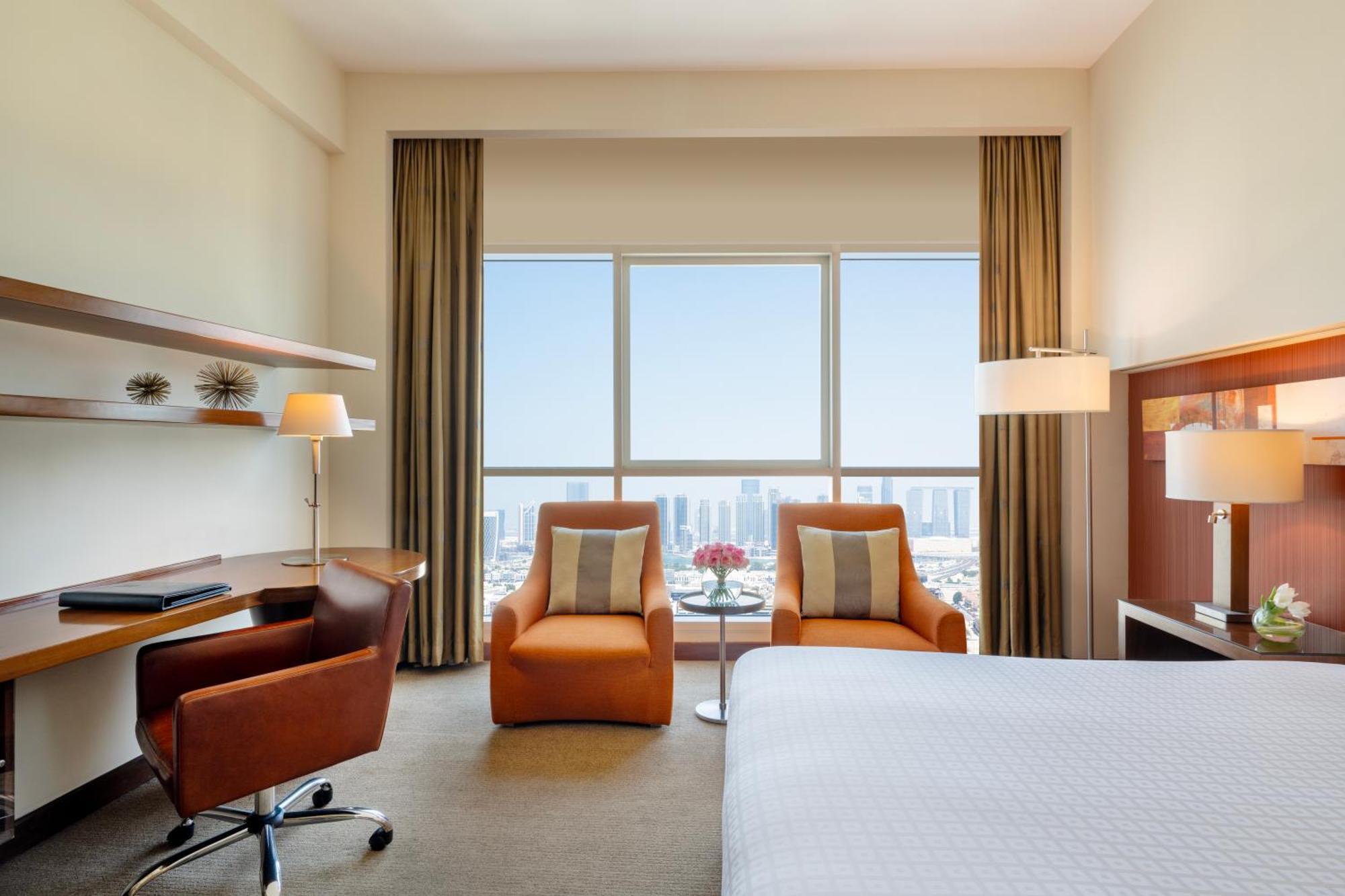 Grand Millennium Al Wahda Hotel And Executive Apartments Abu Dhabi Exterior photo The photo shows a stylish and modern hotel room. It features a comfortable bed with white bedding, a small seating area with two orange chairs and a glass table, and a desk equipped with a lamp and a chair. Large windows provide a view of the outside