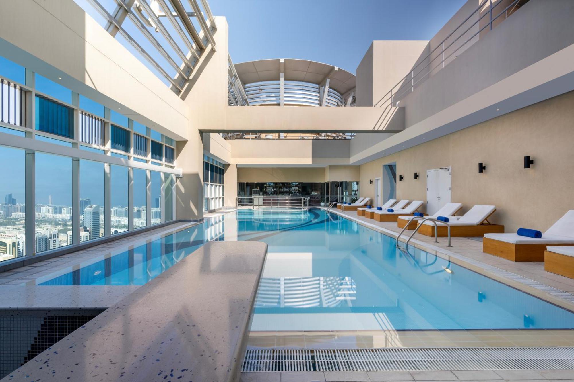 Grand Millennium Al Wahda Hotel And Executive Apartments Abu Dhabi Exterior photo The image shows a modern, luxurious indoor pool area. The pool is large and has a clear, inviting blue water. Surrounding the pool are comfortable lounge chairs arranged neatly. Large windows allow natural light to flood the space, highlighting the s