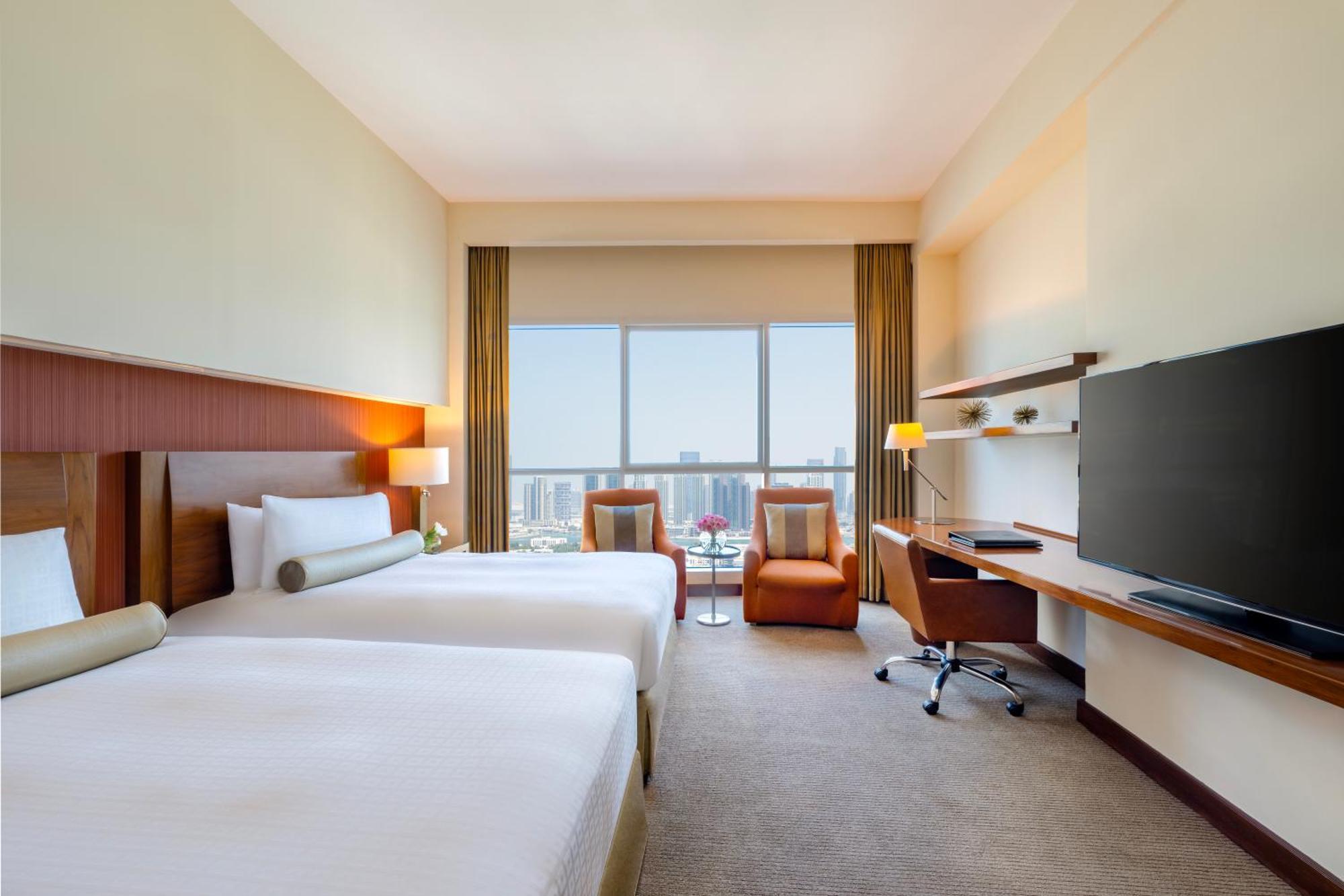 Grand Millennium Al Wahda Hotel And Executive Apartments Abu Dhabi Exterior photo The photo shows a hotel room featuring two beds with white linens. There is a large window with view that allows natural light to fill the space, and it is adorned with light-colored curtains. Opposite the beds, there is a flat-screen television moun