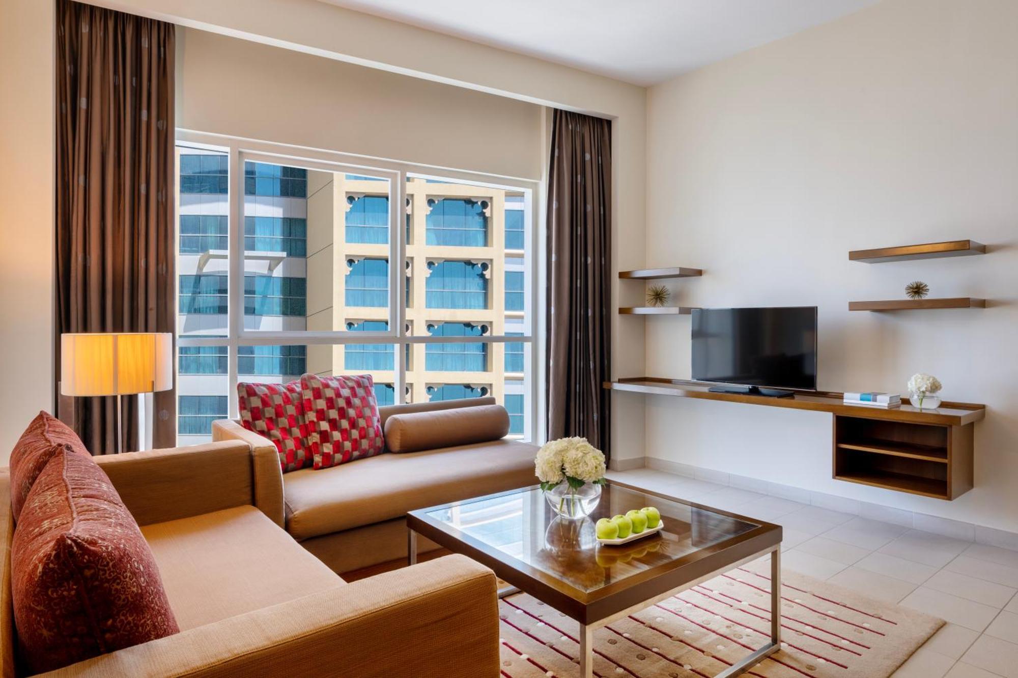 Grand Millennium Al Wahda Hotel And Executive Apartments Abu Dhabi Exterior photo The photo shows a modern living room setup. It features a sofa with decorative cushions, a coffee table with some decorative items on it, and a television mounted on a wall shelf. The room has large windows allowing natural light to enter, and there 