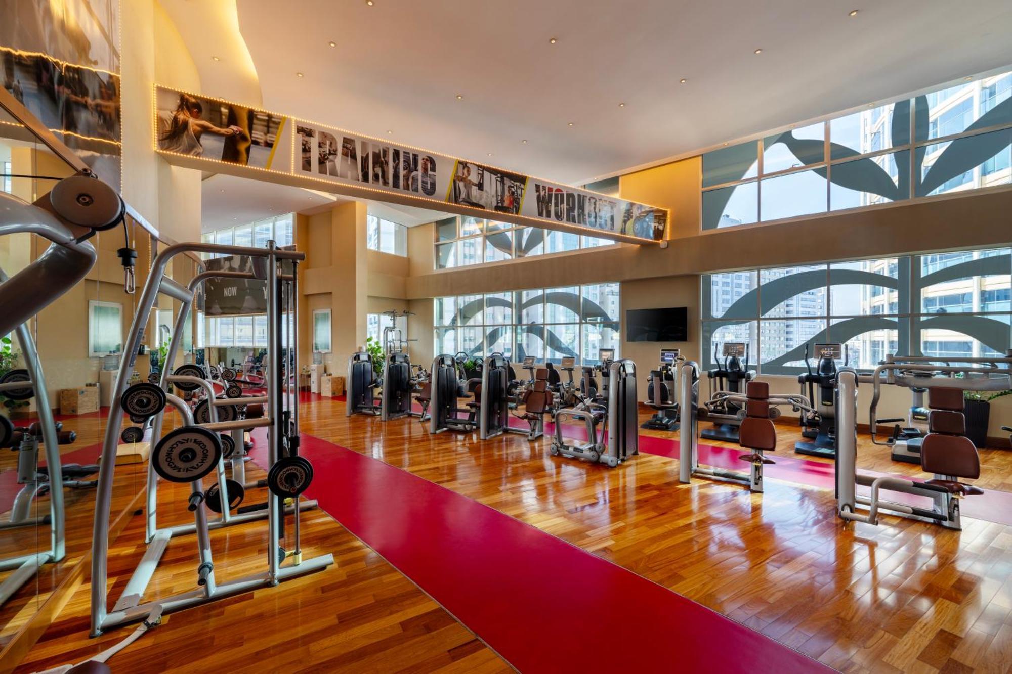 Grand Millennium Al Wahda Hotel And Executive Apartments Abu Dhabi Exterior photo The photo shows a modern gym or fitness center with a spacious and well-lit interior. The flooring is made of polished wood, and there is a prominent red carpet running through the area. The gym is equipped with various exercise machines and weights,