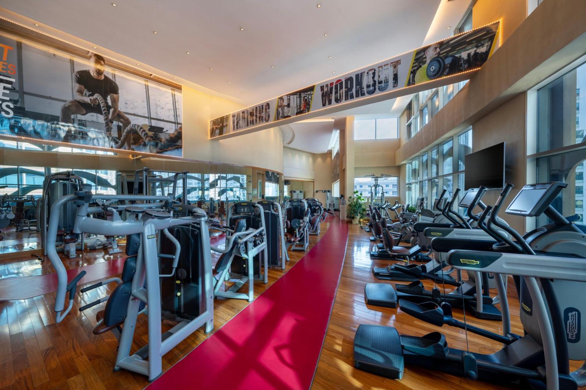 Grand Millennium Al Wahda Hotel And Executive Apartments Abu Dhabi Exterior photo The photo shows a spacious gym interior with a sleek design. There are various exercise machines lined up, including treadmills, elliptical machines, and weight equipment. The floor features polished wood, and a red carpet runs down the center. Large