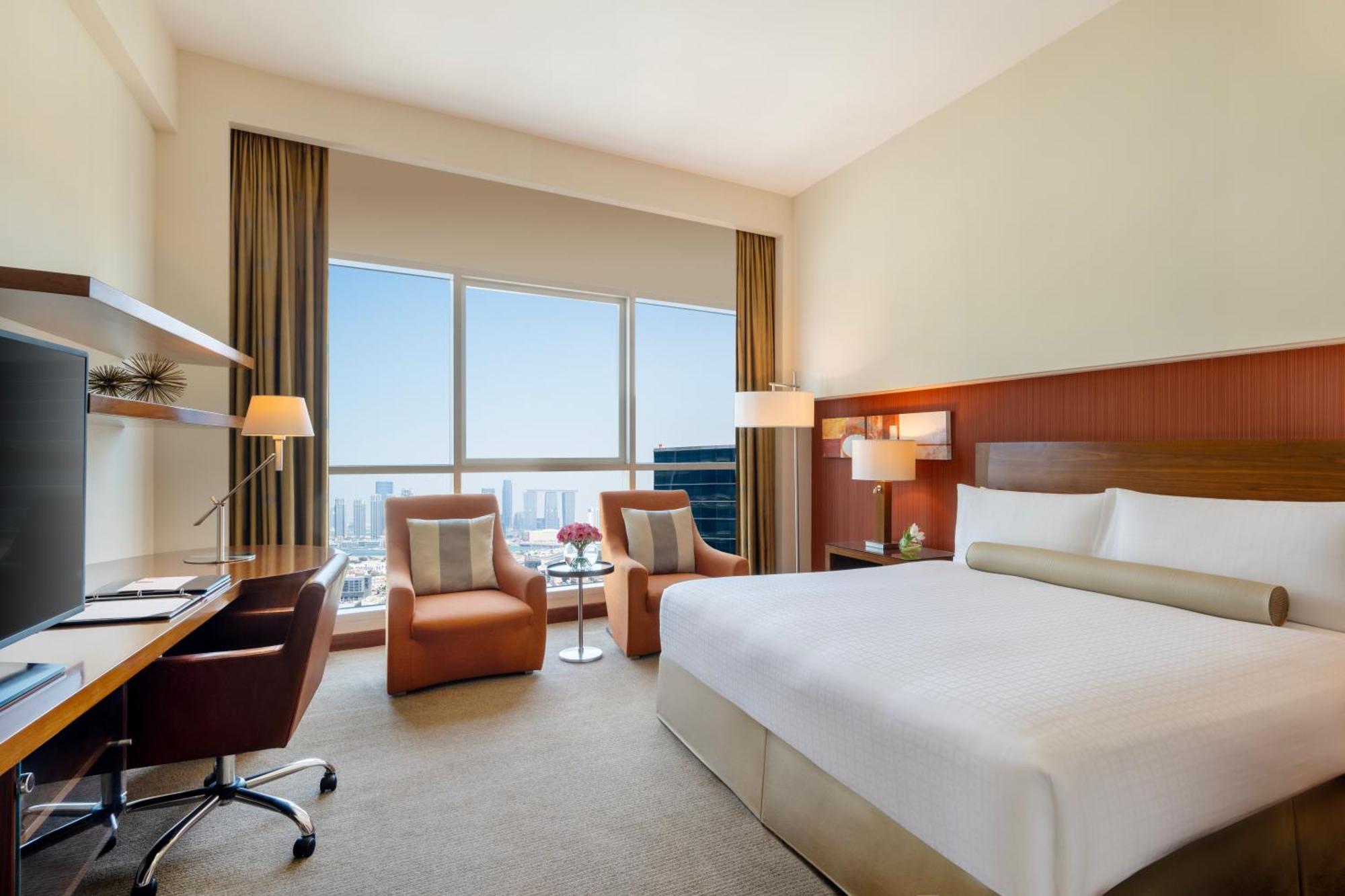 Grand Millennium Al Wahda Hotel And Executive Apartments Abu Dhabi Exterior photo The photo shows a modern hotel room featuring a large bed with white bedding. There are two comfortable chairs positioned near a small table, providing a cozy seating area. A desk with a chair is also present, presumably for work or studying. Large w