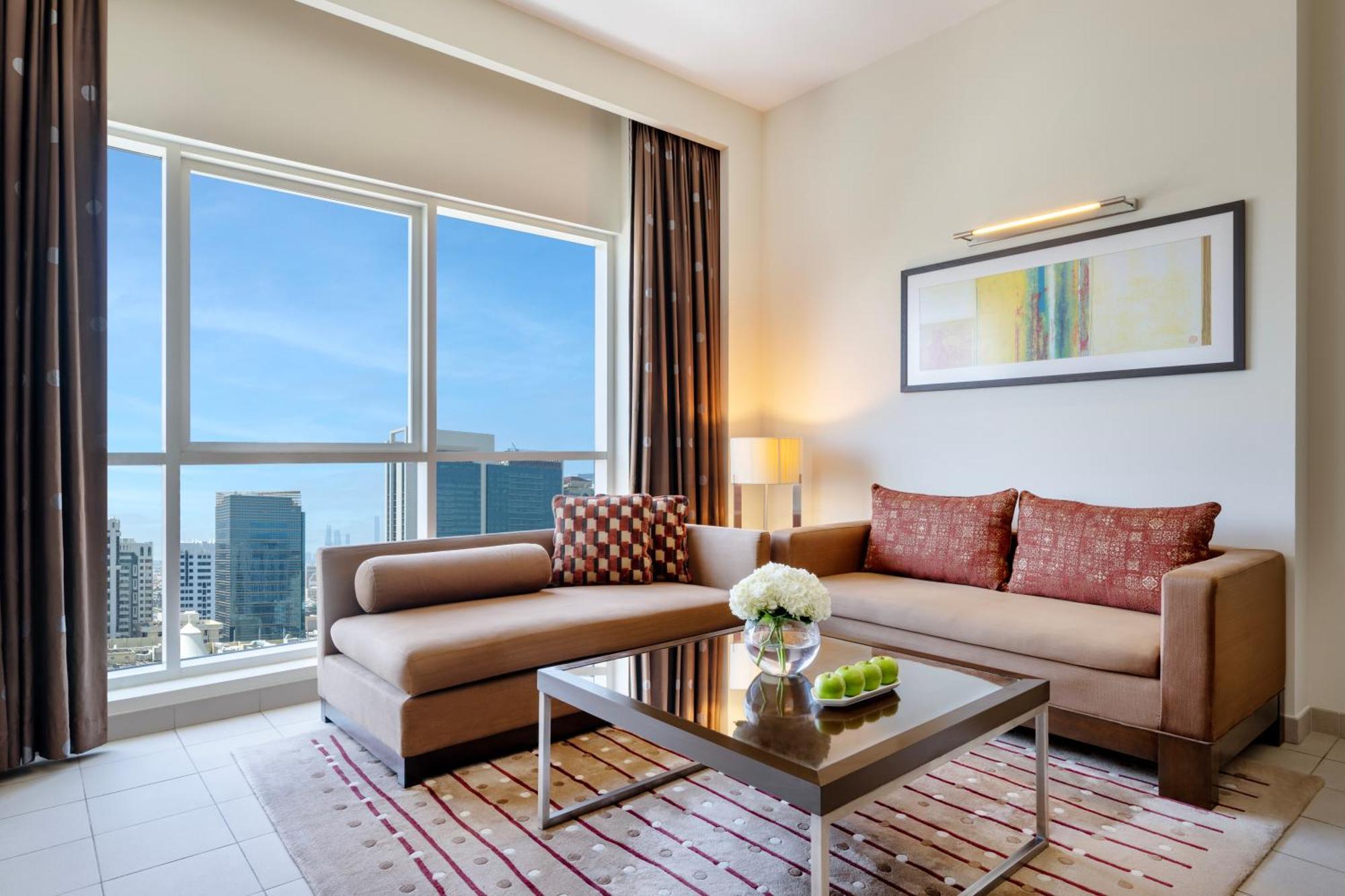 Grand Millennium Al Wahda Hotel And Executive Apartments Abu Dhabi Exterior photo The photo shows a modern living room designed with a contemporary aesthetic. There is a stylish sofa in a light brown color, accented with patterned cushions. A coffee table sits in the center, topped with a decorative arrangement of flowers and a sm