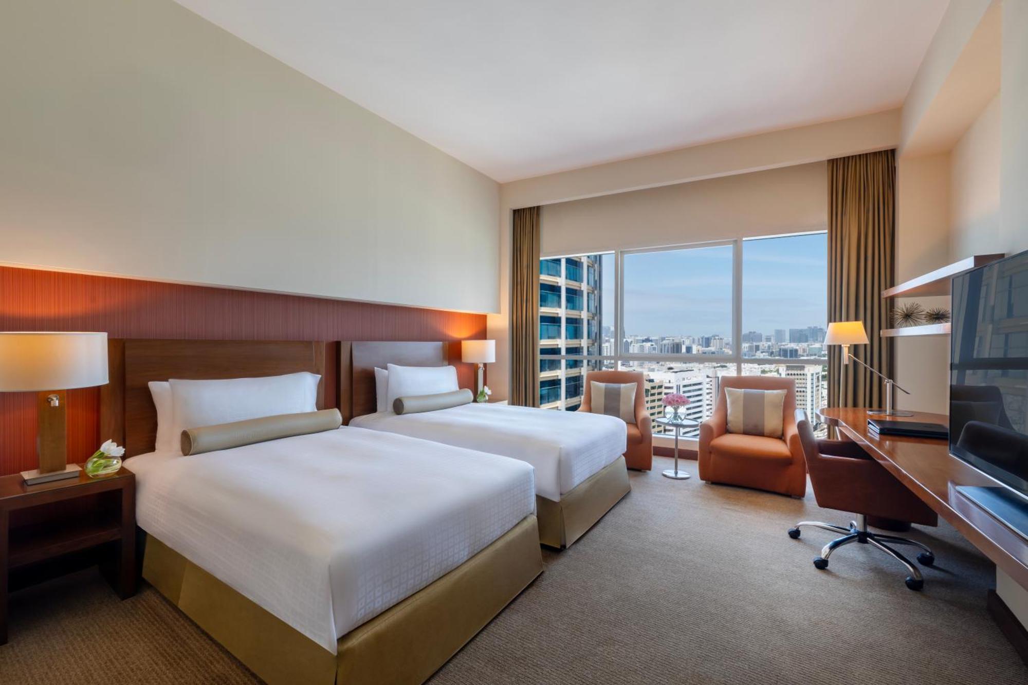 Grand Millennium Al Wahda Hotel And Executive Apartments Abu Dhabi Exterior photo The photo shows a hotel room featuring two twin beds with white linens and decorative pillows. There is a modern desk and chair in one corner, along with a flat-screen TV mounted on the wall. Large windows allow natural light to flood the room, offer