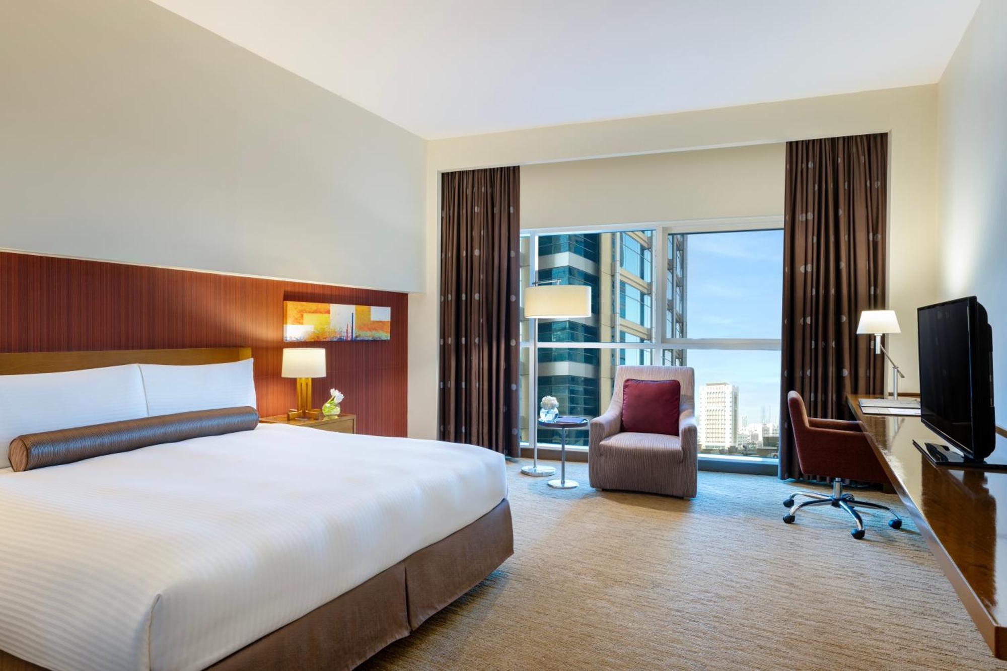 Grand Millennium Al Wahda Hotel And Executive Apartments Abu Dhabi Exterior photo The photo shows a modern hotel room featuring a large bed with white linens and a brown accent. There are bedside tables with lamps on either side of the bed. A comfortable armchair and a small table are positioned near a large window that offers a v