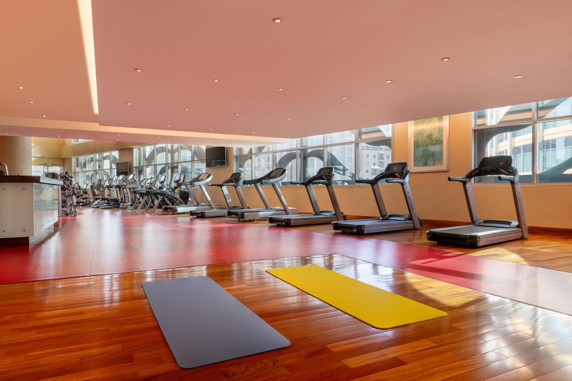 Grand Millennium Al Wahda Hotel And Executive Apartments Abu Dhabi Exterior photo The photo shows a modern gym or fitness area. There are several treadmills lined up against the wall, and the gym features large windows that provide natural light. The flooring is a mix of hardwood and exercise mats, with at least two exercise mats 