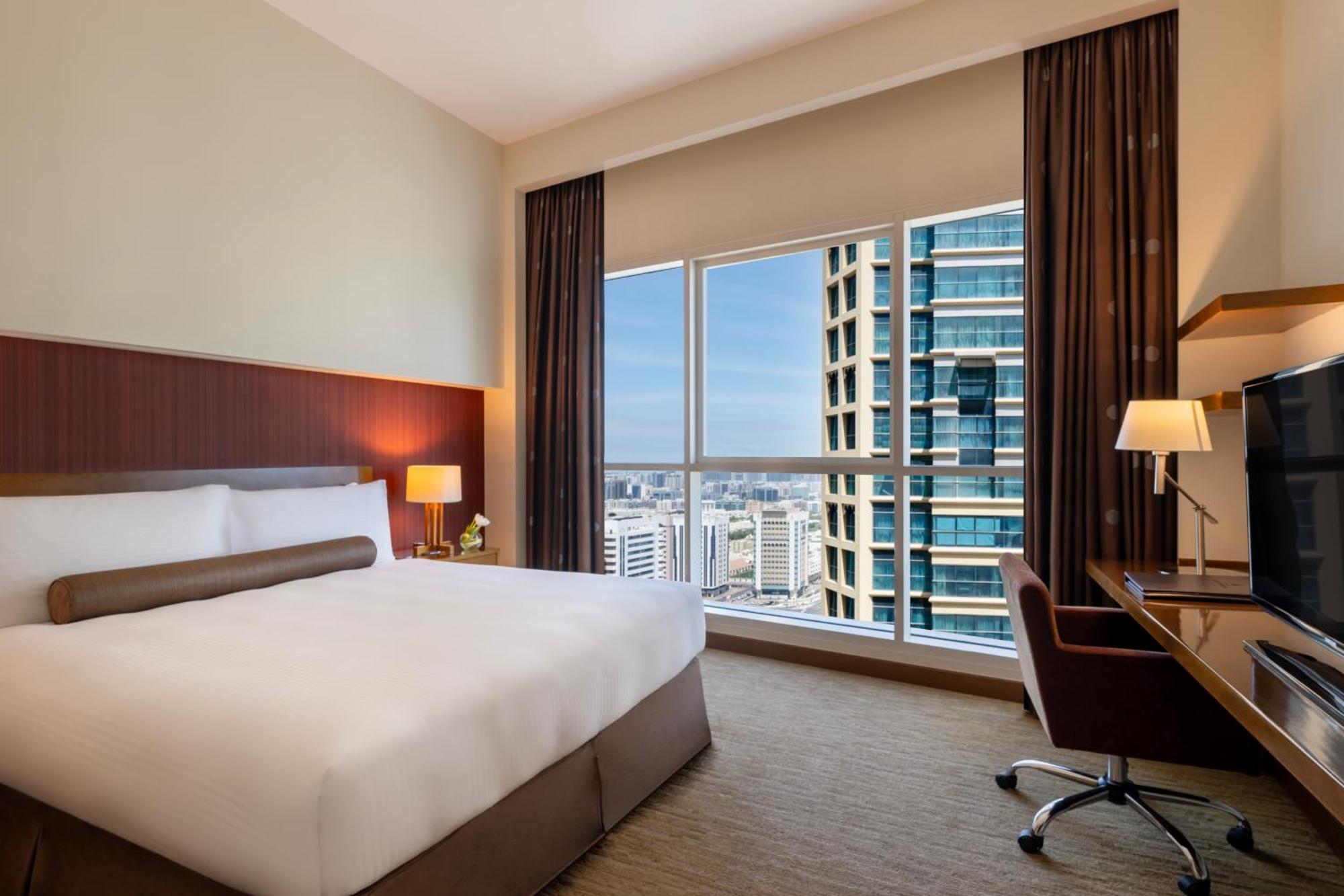 Grand Millennium Al Wahda Hotel And Executive Apartments Abu Dhabi Exterior photo The photo shows a modern hotel room featuring a large bed with a white and beige color scheme. There are bedside lamps on either side of the bed, and a desk with a chair is positioned near a window. The window provides a view of a cityscape with tall