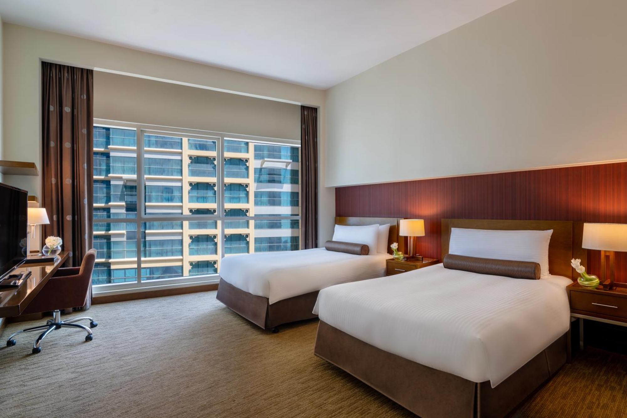 Grand Millennium Al Wahda Hotel And Executive Apartments Abu Dhabi Exterior photo The photo shows a modern hotel room featuring two beds with white linens and brown accents. There are bedside lamps on each side of the beds, and a stylish desk and chair are positioned against one wall. The room has large windows that allow natural 
