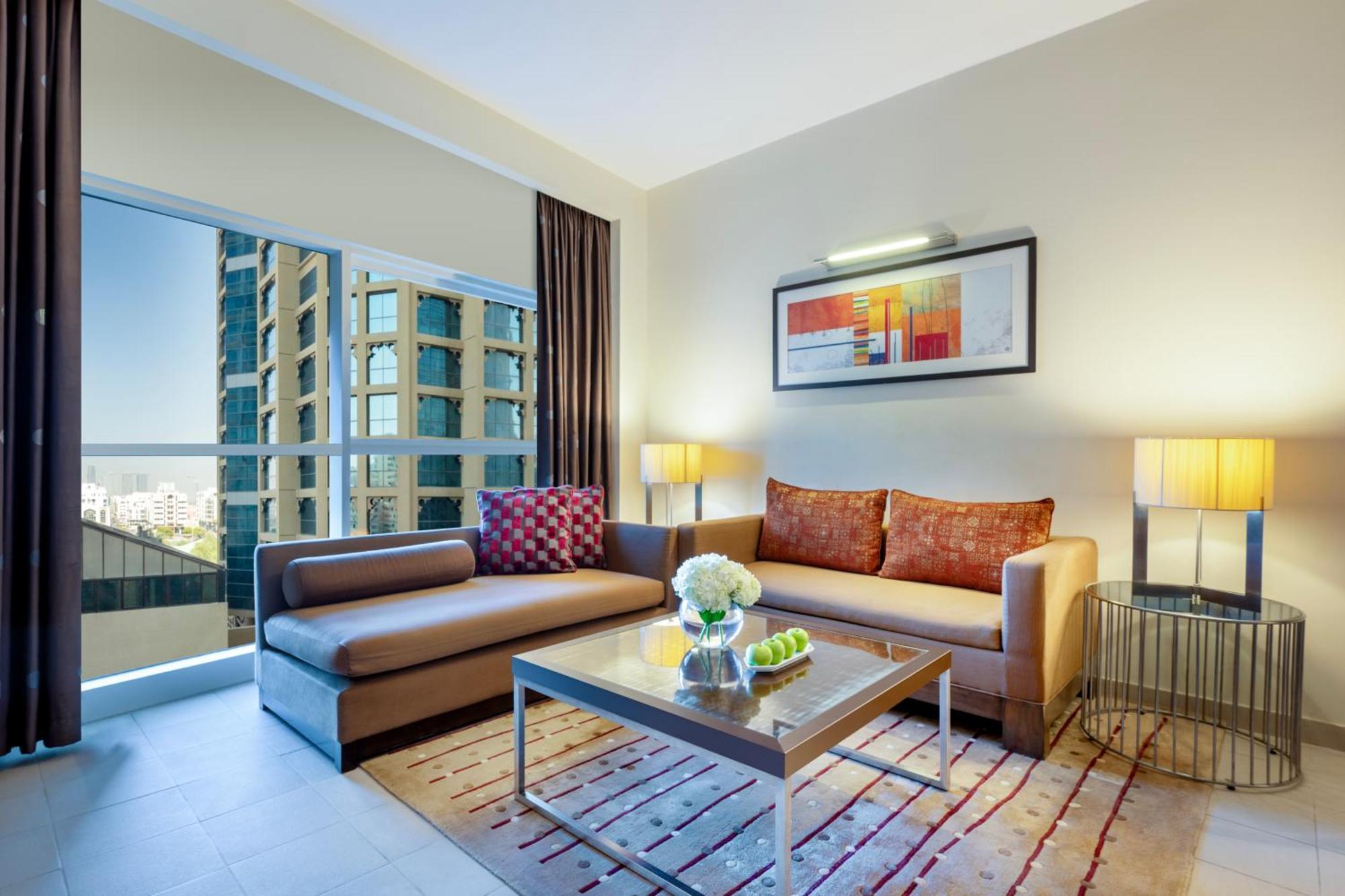 Grand Millennium Al Wahda Hotel And Executive Apartments Abu Dhabi Exterior photo The photo shows a modern and stylish living room. It features a light-colored wall and a large window with a view of a cityscape outside. There is a comfortable brown sofa with decorative pillows, paired with a sleek coffee table. On the table, there