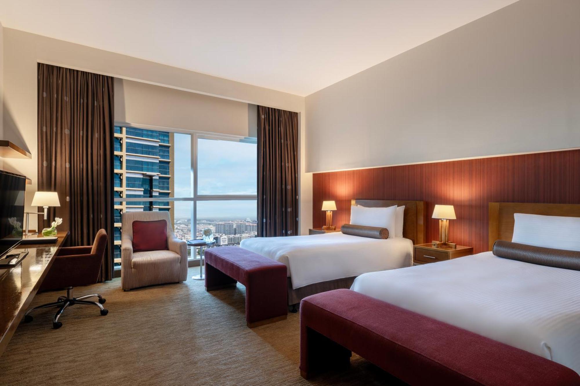Grand Millennium Al Wahda Hotel And Executive Apartments Abu Dhabi Exterior photo The photo shows a modern hotel room featuring two beds, each with a headboard and a small accent pillow. There is a cozy chair in the corner, along with a desk and a lamp. Floor-to-ceiling windows allow natural light to fill the room and offer a view