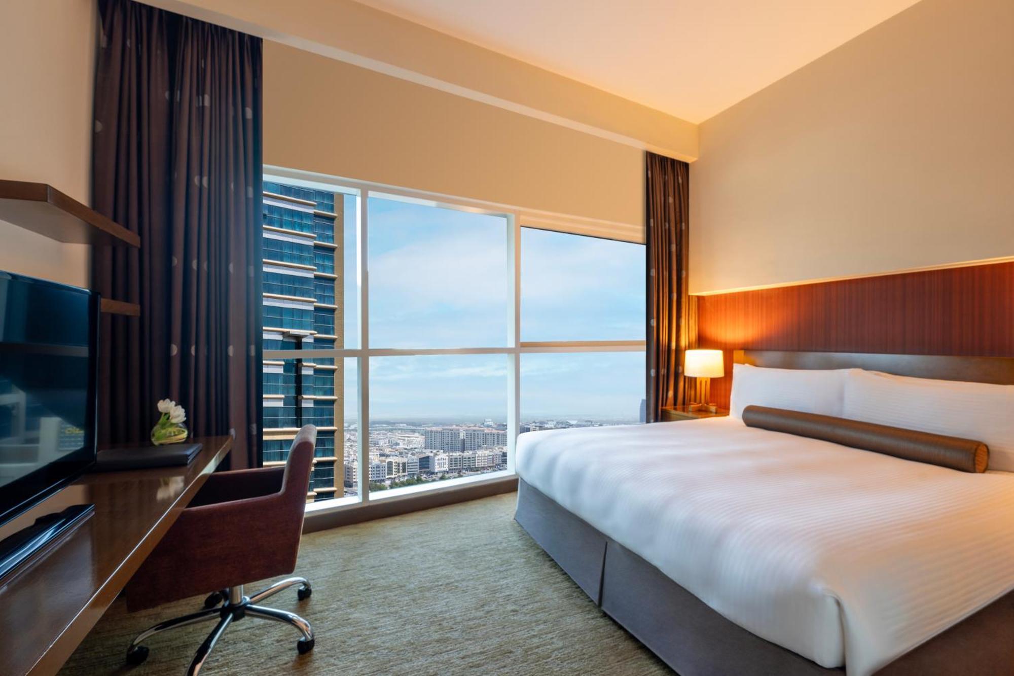 Grand Millennium Al Wahda Hotel And Executive Apartments Abu Dhabi Exterior photo The photo shows a modern hotel room featuring a large bed with white linens and a decorative throw pillow. There is a stylish desk and chair situated beside a window that offers a view of the cityscape outside. The room has large windows allowing nat