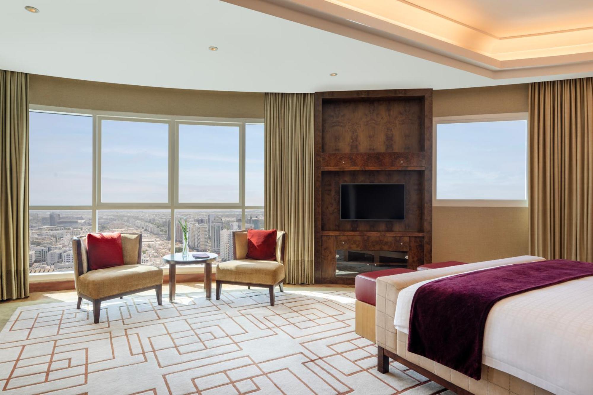 Grand Millennium Al Wahda Hotel And Executive Apartments Abu Dhabi Exterior photo The photo shows a luxurious hotel room with a modern design. It features large windows that provide an expansive view of the city. The room includes a comfortable bed with elegant bedding, and there are two armchairs with decorative cushions near a s