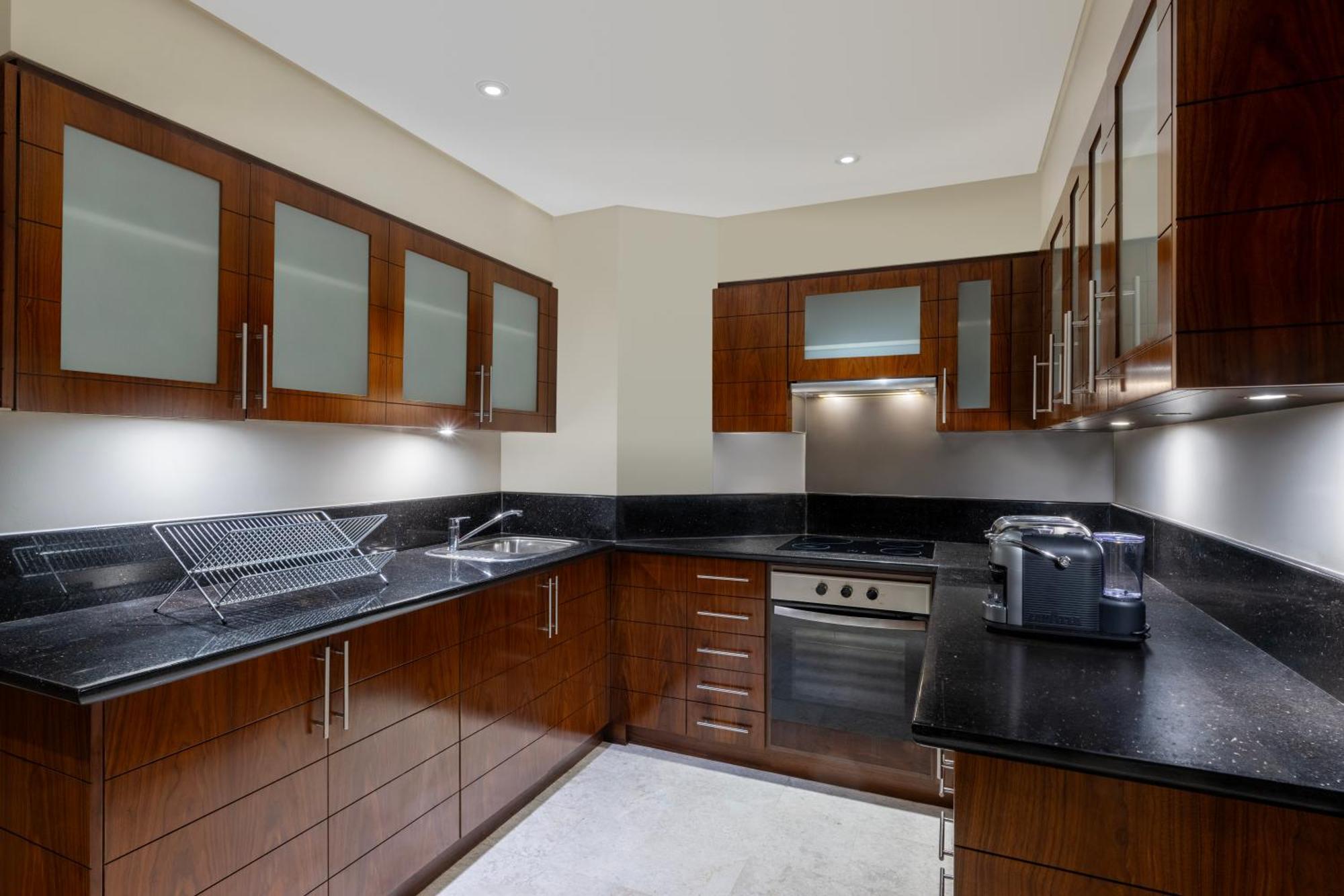 Grand Millennium Al Wahda Hotel And Executive Apartments Abu Dhabi Exterior photo The photo shows a modern kitchen. It features dark wood cabinetry with glass panels on some of the doors. The countertops are made of black stone, and there is a stainless steel sink with a drying rack next to it. The kitchen includes a built-in oven