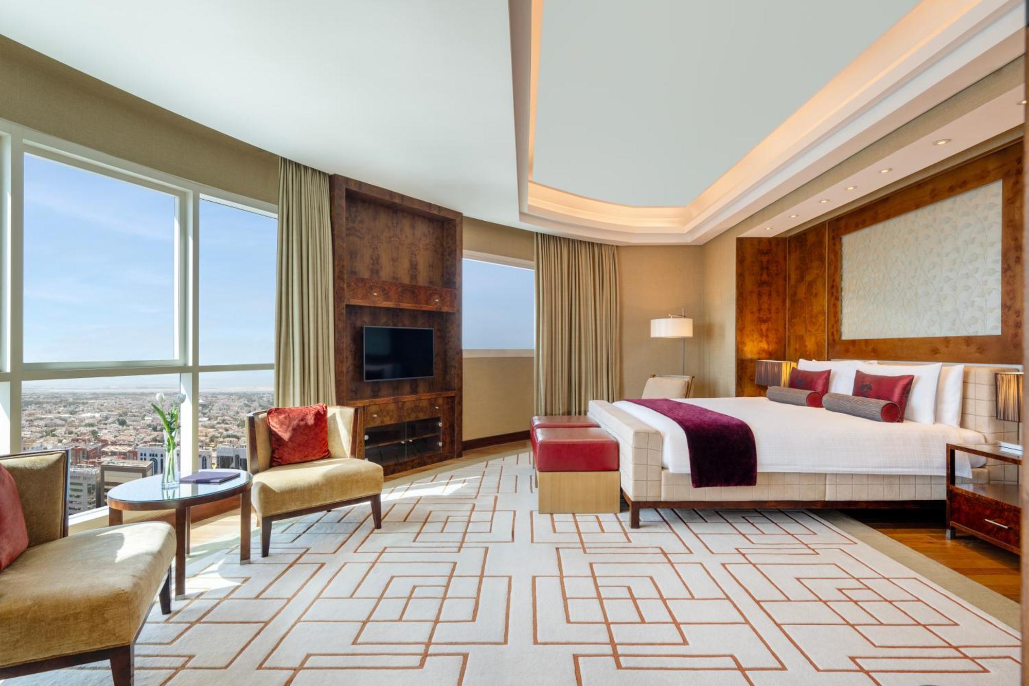 Grand Millennium Al Wahda Hotel And Executive Apartments Abu Dhabi Exterior photo The photo showcases a luxurious hotel room featuring a modern design. It includes a large bed with decorative pillows, a stylish armchair and a small table, and a bench at the foot of the bed. There are large windows providing a panoramic view of the