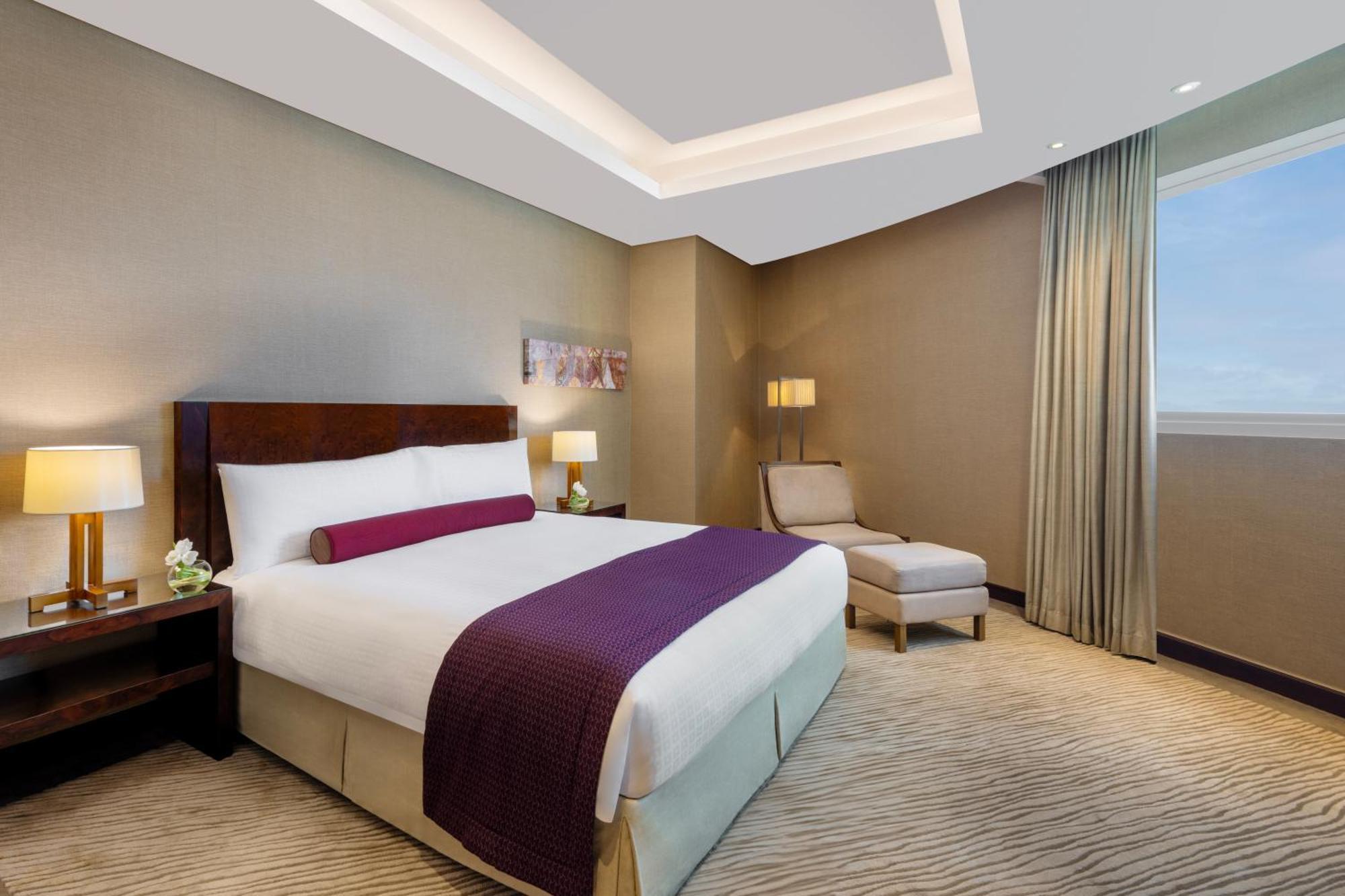 Grand Millennium Al Wahda Hotel And Executive Apartments Abu Dhabi Exterior photo The photo shows a modern hotel room featuring a bed with a white comforter and a purple accent pillow. Next to the bed, there is a small bedside table with a lamp. A comfortable chaise lounge chair is positioned nearby. The room has soft lighting, an