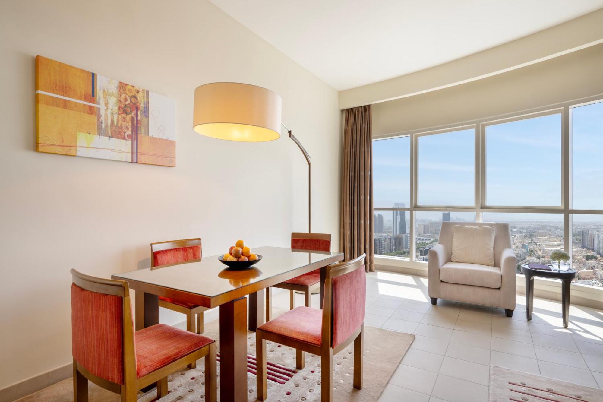Grand Millennium Al Wahda Hotel And Executive Apartments Abu Dhabi Exterior photo The photo shows a well-lit dining area featuring a modern table with four chairs. The table has a bowl of fruit in the center. To the left, there is a standing lamp with a large shade. Behind the dining area, large windows allow natural light to fill