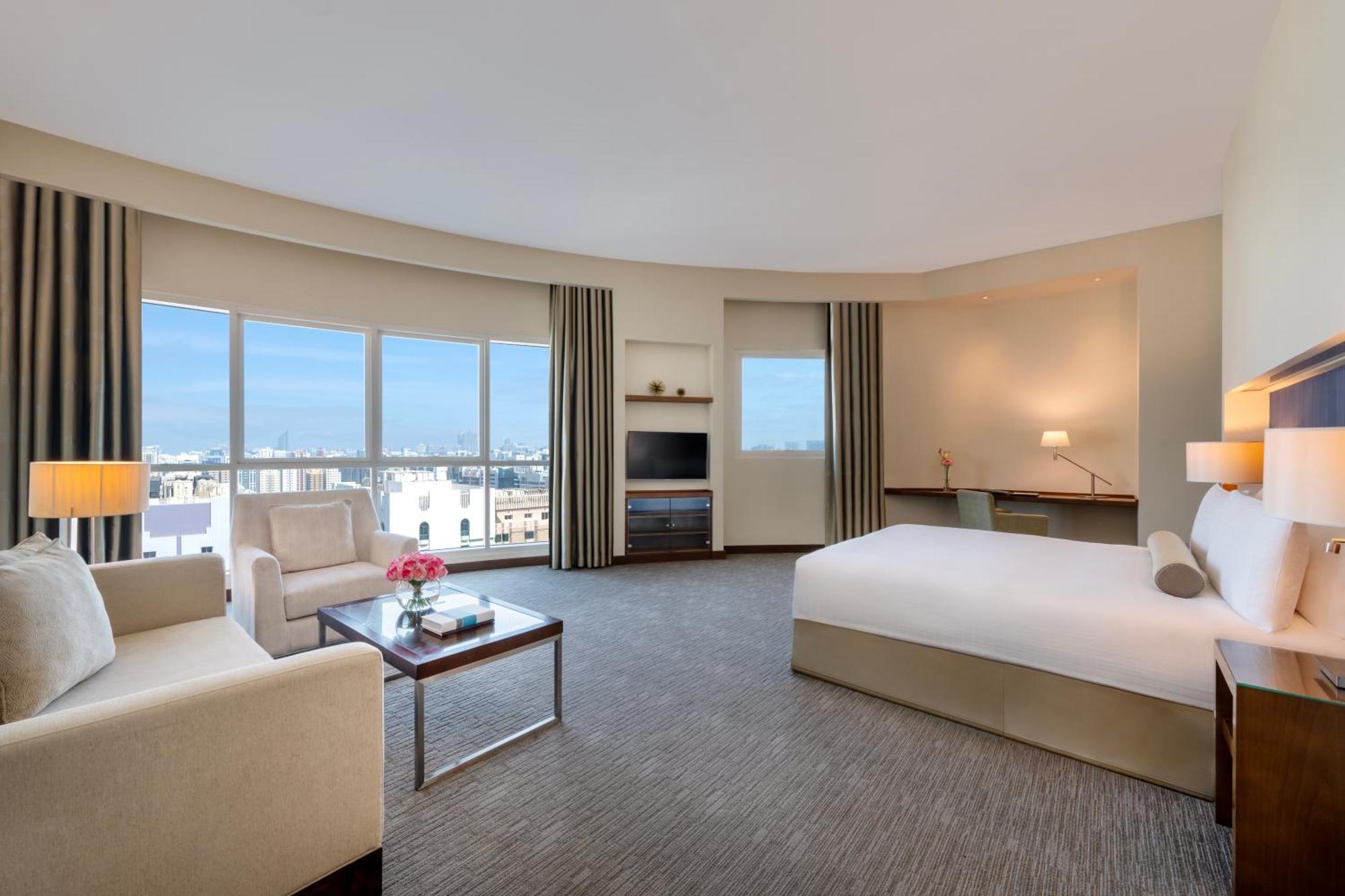 Grand Millennium Al Wahda Hotel And Executive Apartments Abu Dhabi Exterior photo The photo shows a modern hotel room featuring a large, comfortable bed with white linens. There is a cozy seating area with a light-colored sofa and a small table that has a floral arrangement on it. Large windows allow natural light to fill the spac