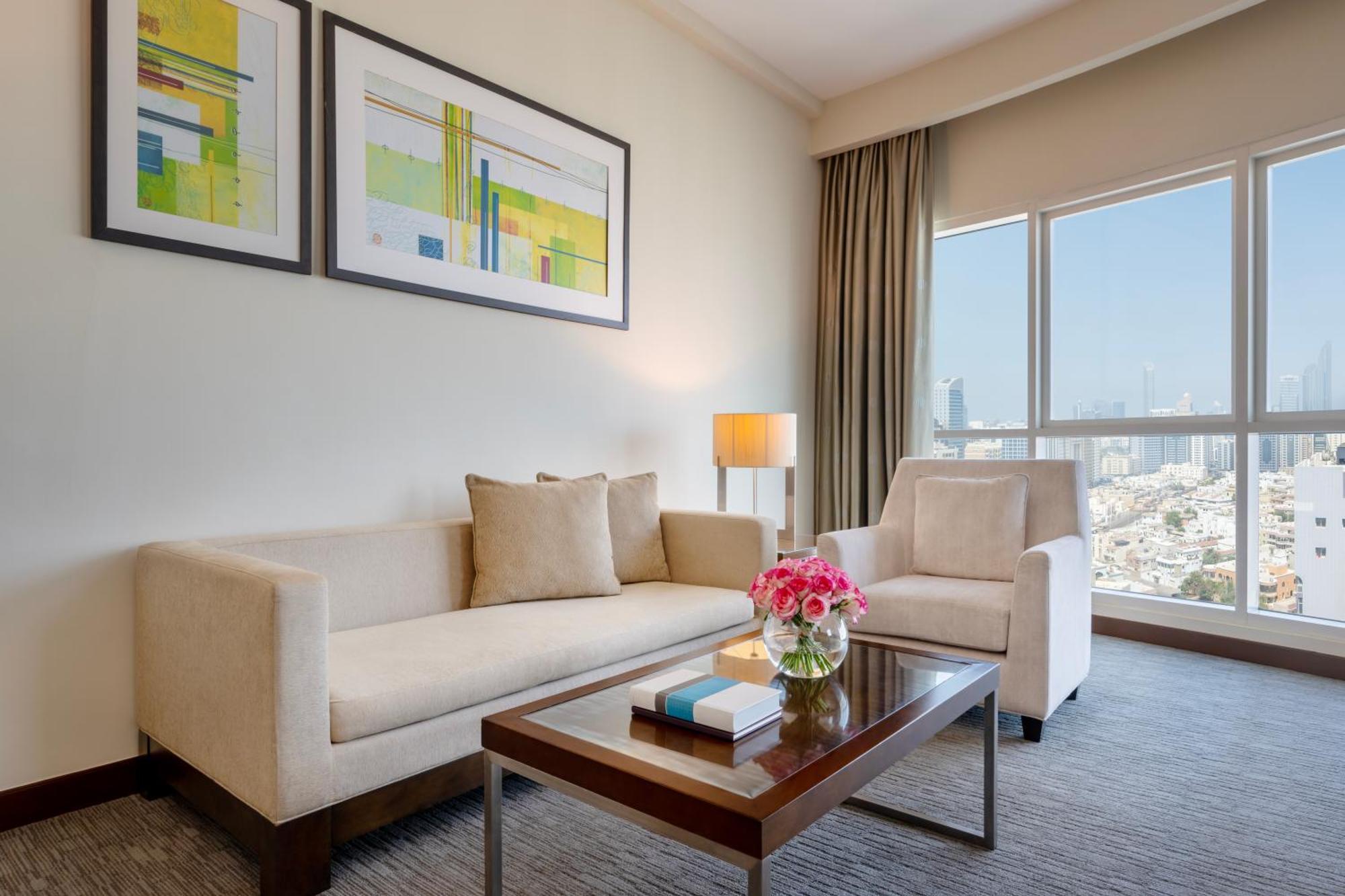 Grand Millennium Al Wahda Hotel And Executive Apartments Abu Dhabi Exterior photo The photo shows a stylish and modern living area. It features a light-colored sofa and an armchair, both positioned near a large window that offers a view of the outside cityscape. A coffee table sits in the middle, adorned with a small bouquet of fl