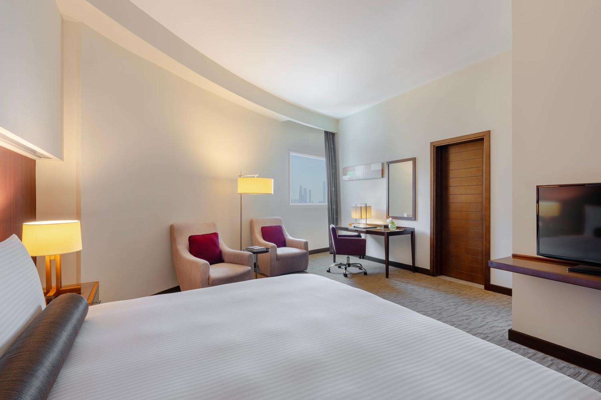 Grand Millennium Al Wahda Hotel And Executive Apartments Abu Dhabi Exterior photo The photo shows a modern hotel room with a clean and spacious layout. In the foreground, there is a large bed with a white duvet. To one side, there are two comfortable-looking armchairs in a light color, each with a decorative pillow. A lamp stands 