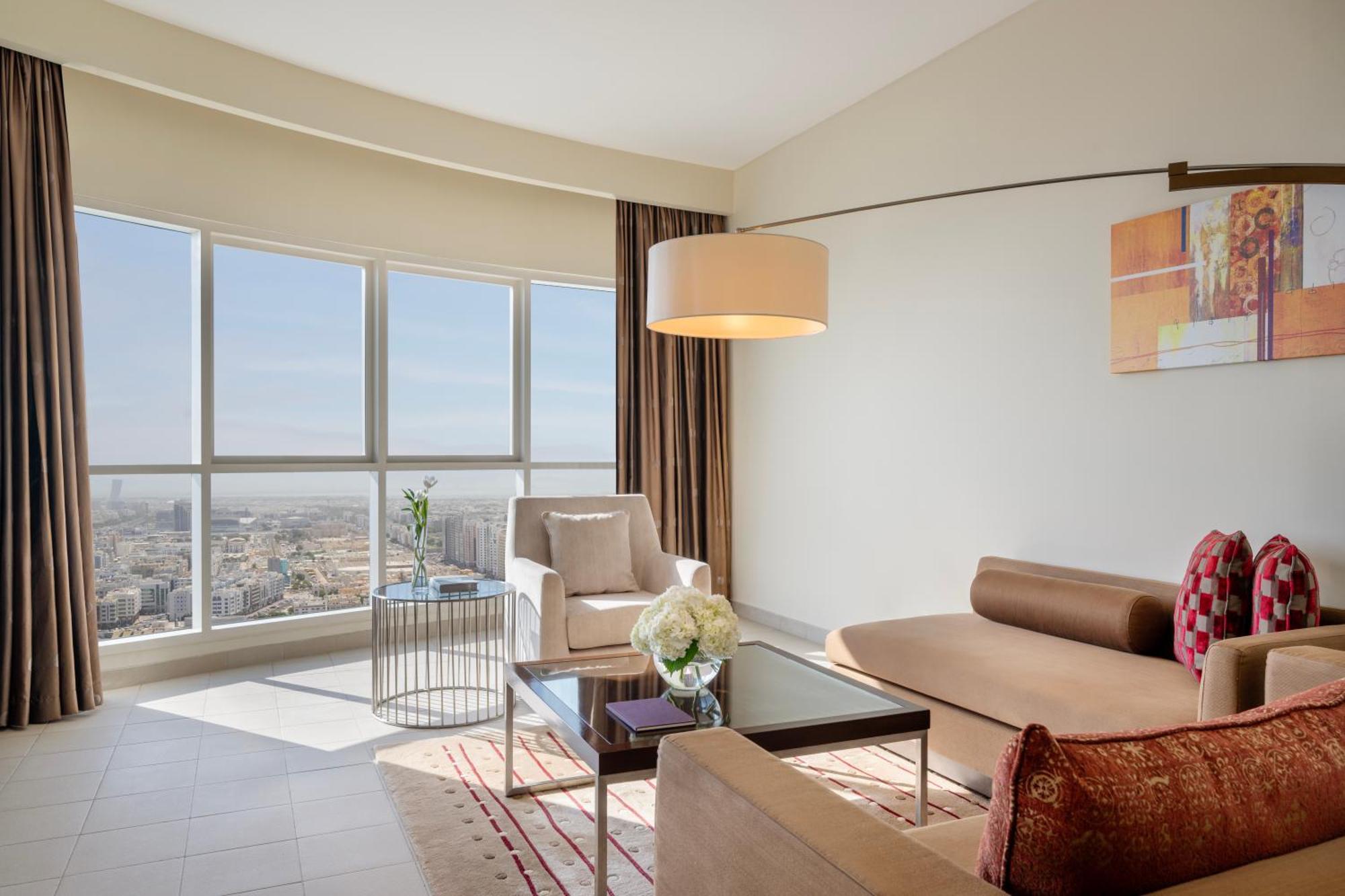 Grand Millennium Al Wahda Hotel And Executive Apartments Abu Dhabi Exterior photo The photo depicts a modern and stylish living room. It features large windows that allow ample natural light, showing a scenic view outside. The space includes a comfortable seating area with an upholstered couch and armchairs. A coffee table is plac