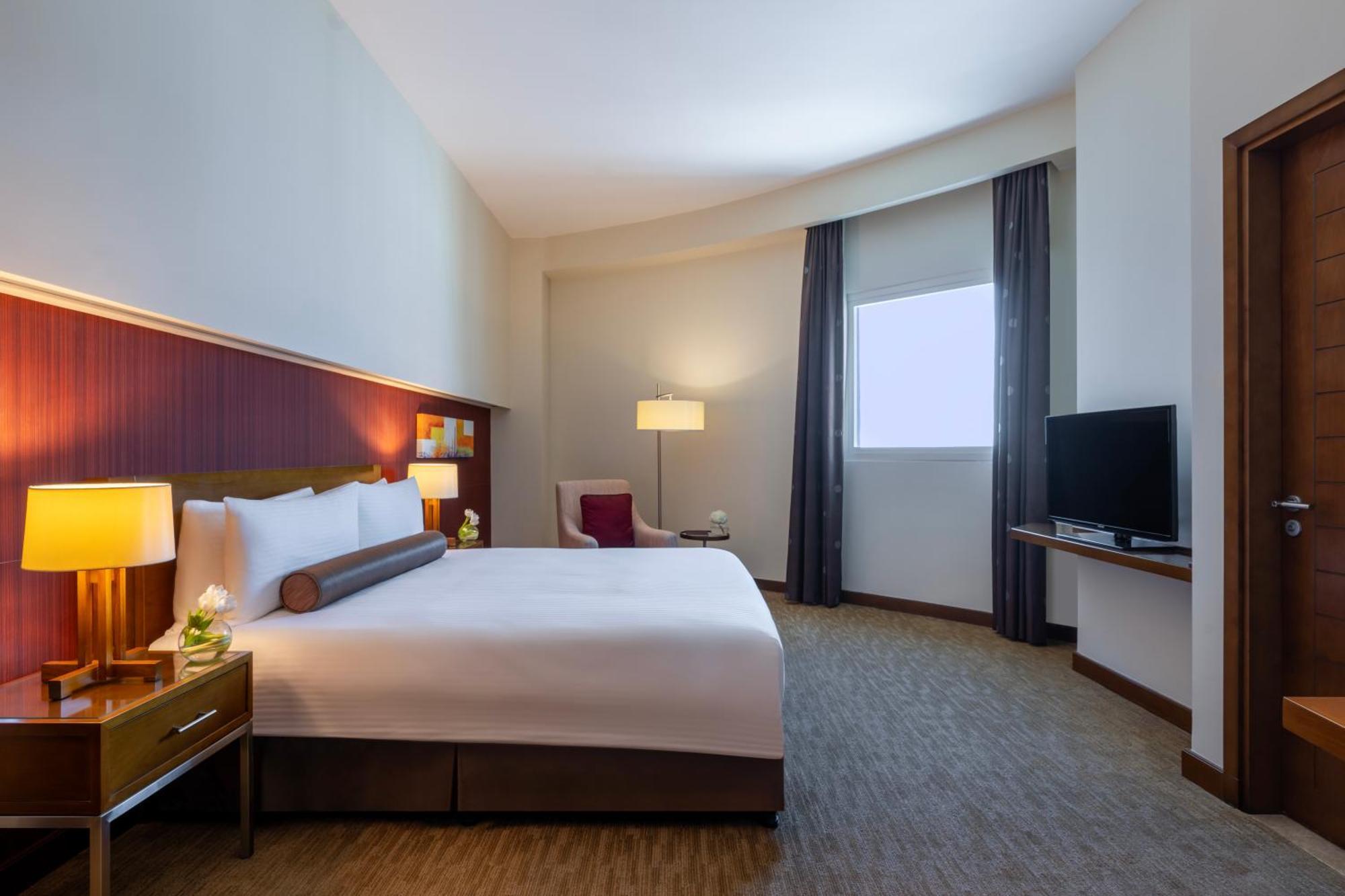 Grand Millennium Al Wahda Hotel And Executive Apartments Abu Dhabi Exterior photo The image shows a modern hotel room. It features a large bed with white linens and a decorative bolster pillow. There are two bedside lamps on either side of the bed and a small seating area with a chair and a side table. A flat-screen TV is mounted 