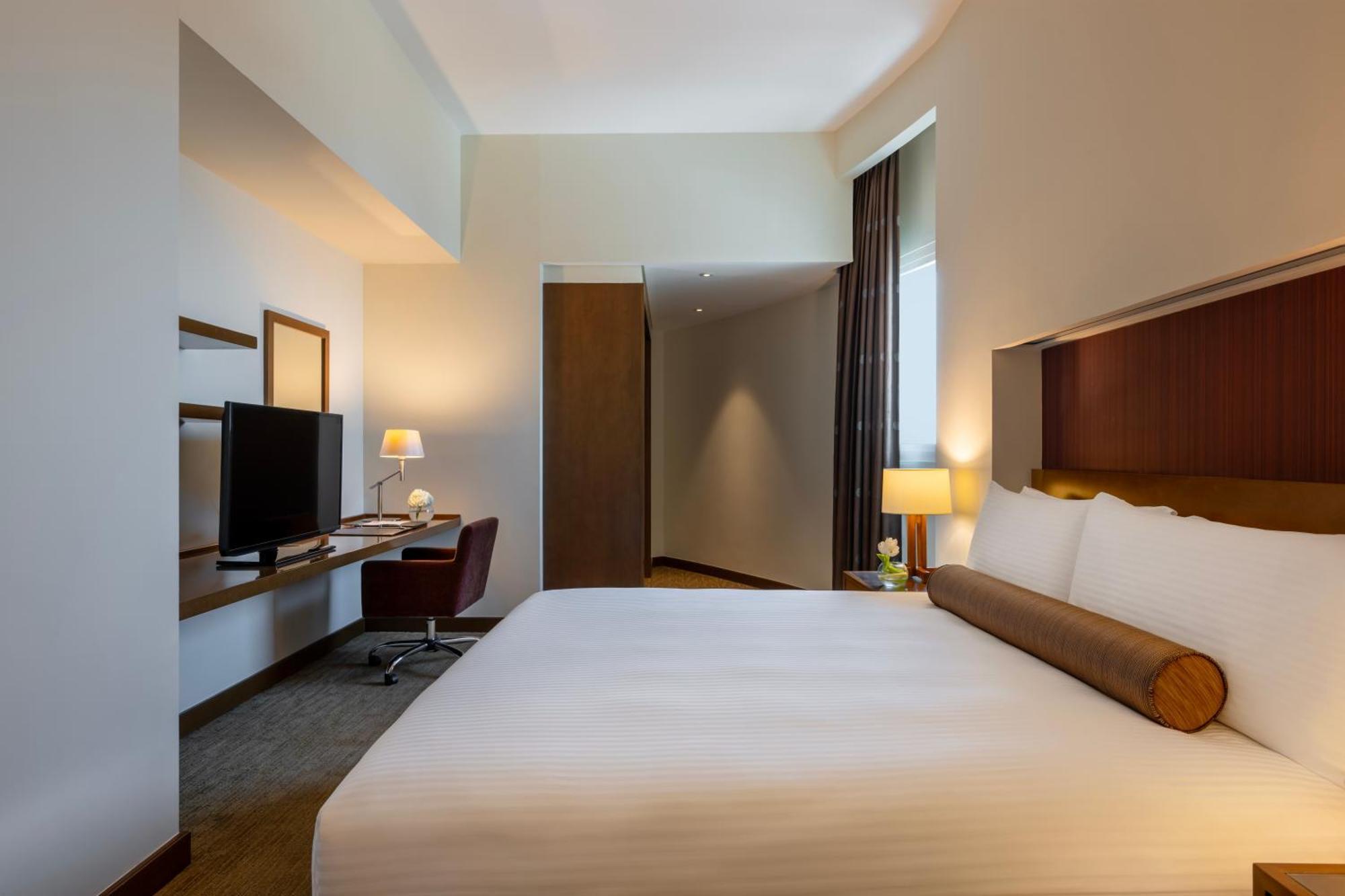 Grand Millennium Al Wahda Hotel And Executive Apartments Abu Dhabi Exterior photo The photo shows a modern hotel room featuring a queen-sized bed with white linens and a decorative bolster pillow. To the left, there is a wooden desk with a comfortable chair, a desktop computer or TV, and a lamp. The room has neutral-colored walls 