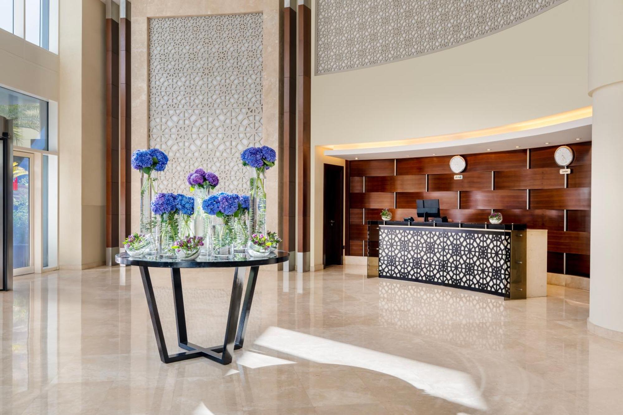 Grand Millennium Al Wahda Hotel And Executive Apartments Abu Dhabi Exterior photo The photo shows a modern hotel lobby. In the foreground, there is a round table with elegant blue flowers in glass vases, complementing the sophisticated decor. Behind the table is a reception desk featuring a decorative, patterned front, likely made