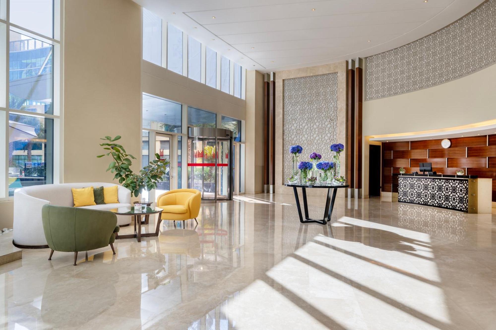 Grand Millennium Al Wahda Hotel And Executive Apartments Abu Dhabi Exterior photo The image shows a modern lobby area with a sleek design. There are large windows allowing natural light to fill the space. In the center, there's a circular table with decorative blue flowers. To one side, there are two armchairs—one yellow and one g