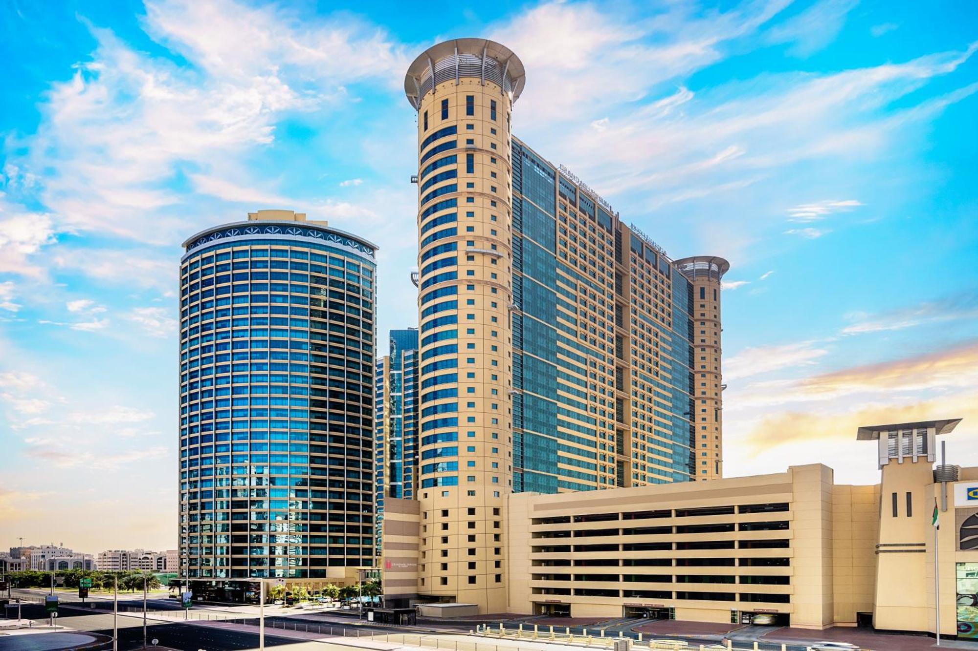 Grand Millennium Al Wahda Hotel And Executive Apartments Abu Dhabi Exterior photo The photo depicts a modern architectural landscape featuring two prominent high-rise buildings and a parking structure. The buildings exhibit a mix of glass and stone facades, showcasing contemporary design elements. The structure on the left is roun