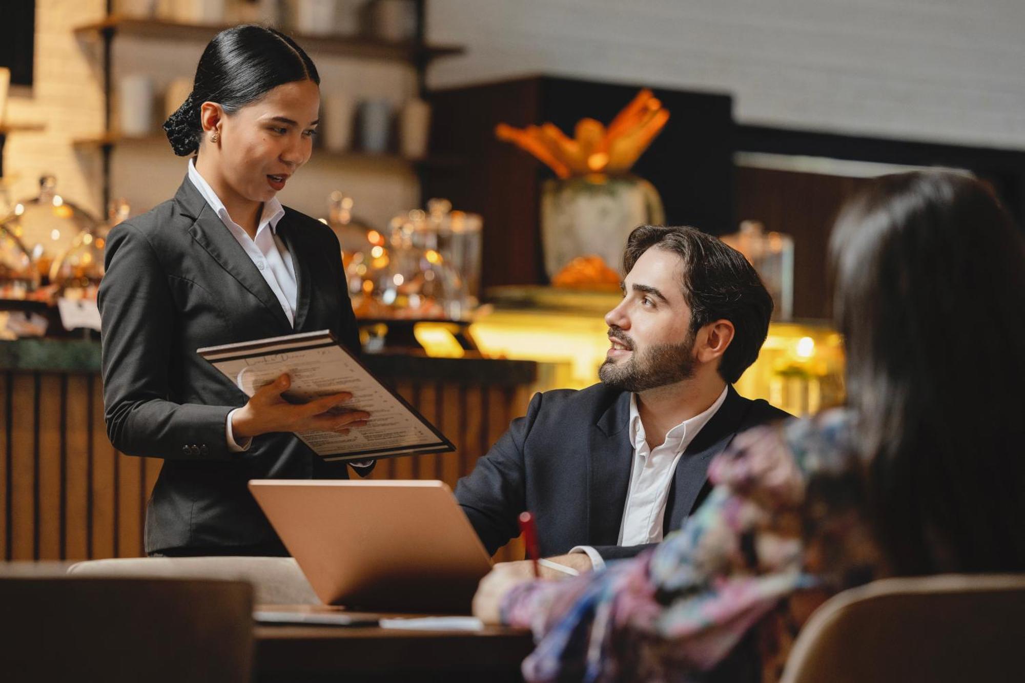 Grand Millennium Al Wahda Hotel And Executive Apartments Abu Dhabi Exterior photo The photo shows a scene in a restaurant or cafe where a female server is presenting a menu to a male customer seated at a table. The man, dressed in a suit, is looking up at her with interest while sitting in front of a laptop. To his side, there is 