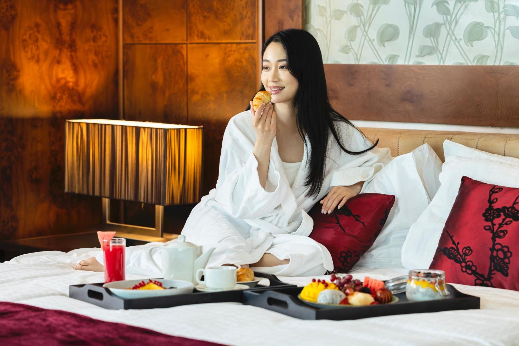 Grand Millennium Al Wahda Hotel And Executive Apartments Abu Dhabi Exterior photo The photo shows a woman sitting on a bed, wearing a white robe. She has long black hair and is smiling while eating a pastry. There are two trays on the bed filled with breakfast items, including fruits, pastries, a pot of tea, and a drink, likely ju