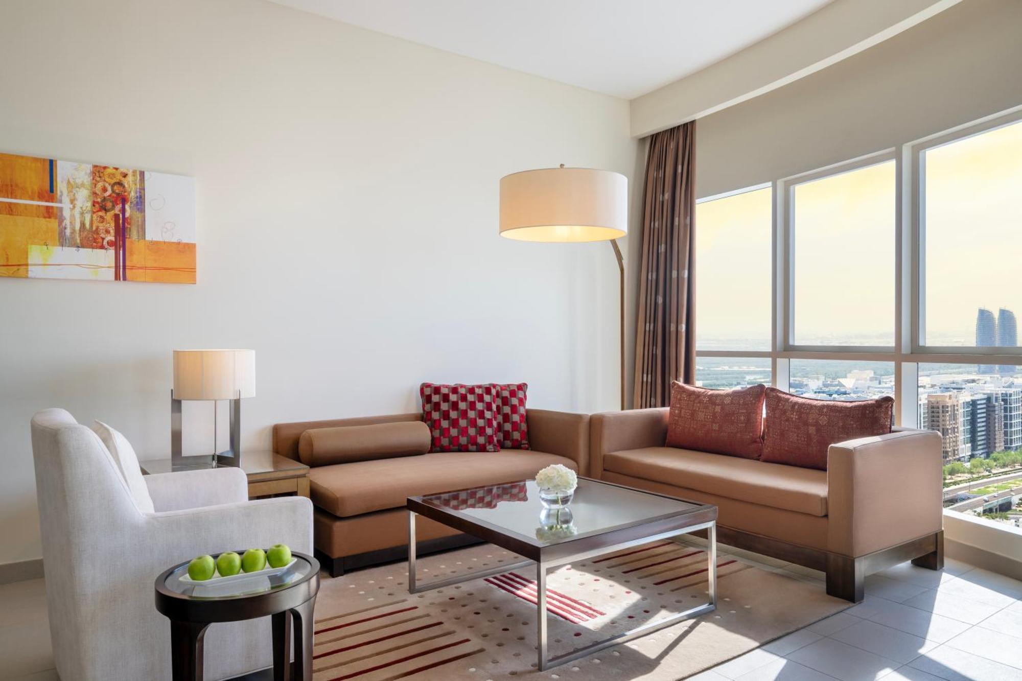 Grand Millennium Al Wahda Hotel And Executive Apartments Abu Dhabi Exterior photo The photo shows a modern living room with large windows allowing natural light to enter. There are two sofas: one is a light-colored, plush armchair, and the other is a brown sectional sofa with decorative cushions. A sleek glass coffee table is in t