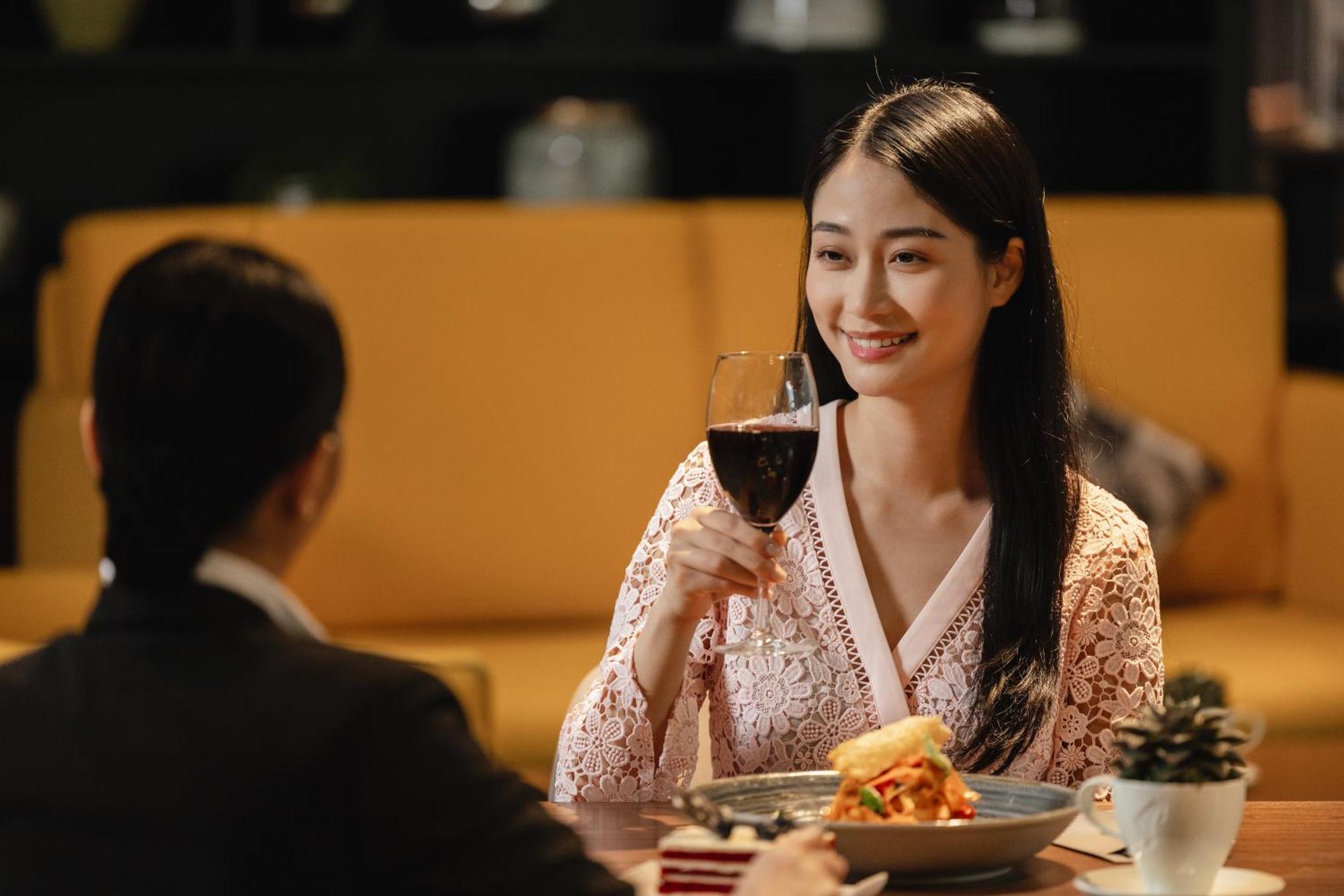 Grand Millennium Al Wahda Hotel And Executive Apartments Abu Dhabi Exterior photo The photo depicts a dining scene where a woman is sitting at a table, smiling and holding a glass of red wine. She appears to be engaged in conversation with another person, who is partially visible. The setting is cozy and elegant, with a rich color