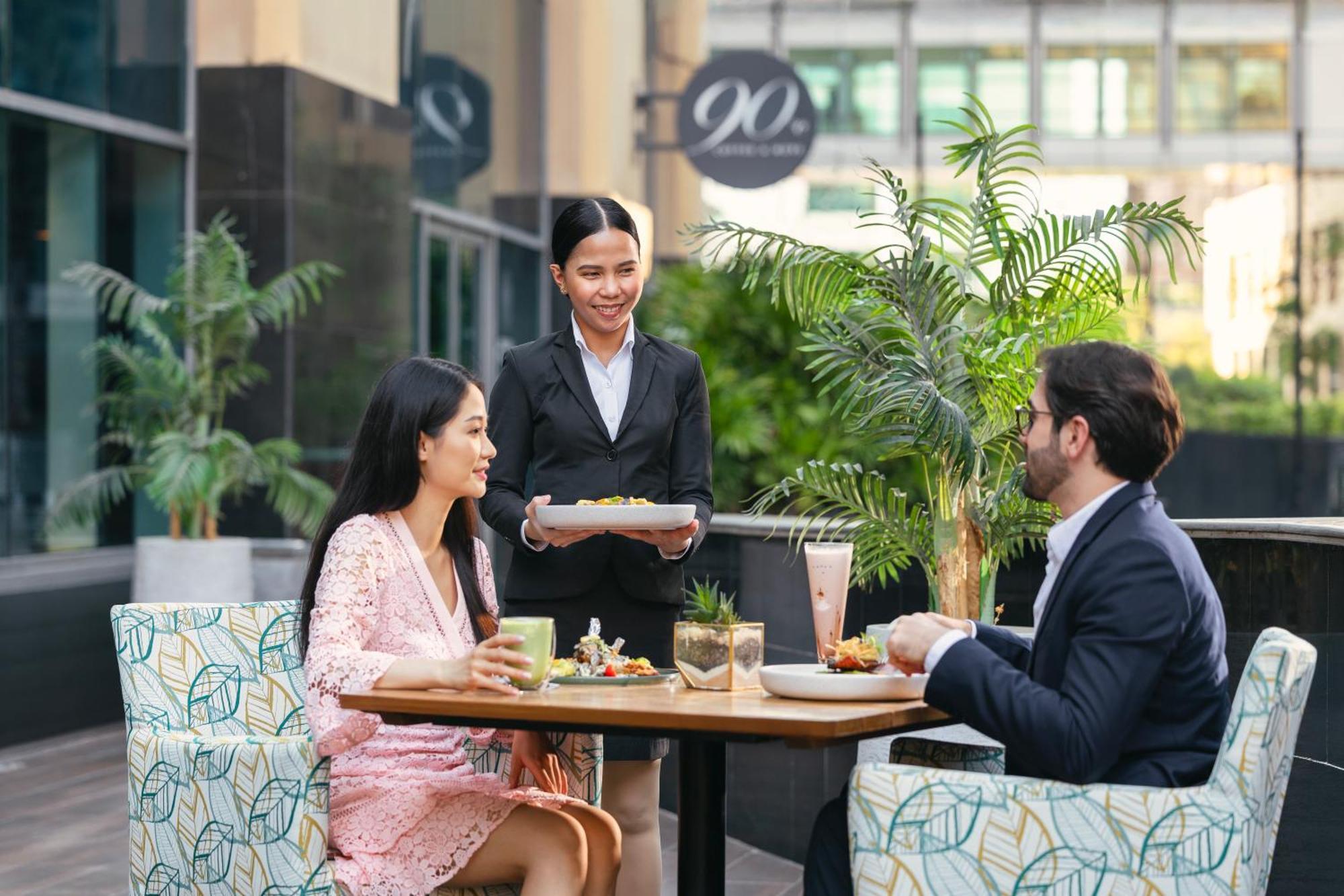 Grand Millennium Al Wahda Hotel And Executive Apartments Abu Dhabi Exterior photo The photo depicts a dining scene at an outdoor establishment. A young woman is seated at a table, enjoying a drink, while a man across from her is eating a meal. A waitress, dressed in a black suit, is serving food to them. The setting includes leafy