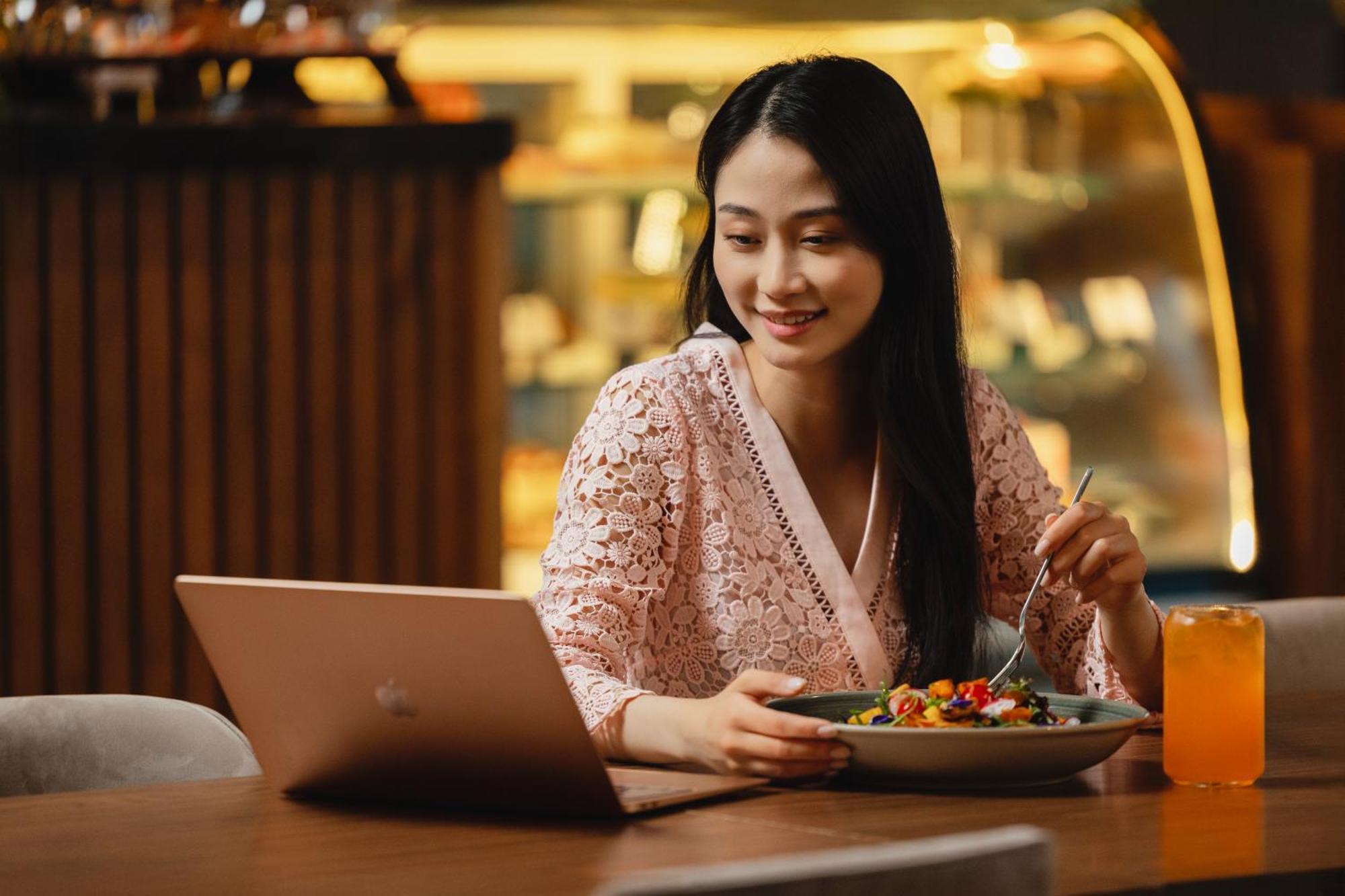 Grand Millennium Al Wahda Hotel And Executive Apartments Abu Dhabi Exterior photo The photo shows a woman sitting at a table in a cafe or restaurant. She is dressed in a light, patterned outfit and is smiling as she looks at a laptop in front of her. On the table, she has a plate of colorful food and is holding a fork, suggesting 