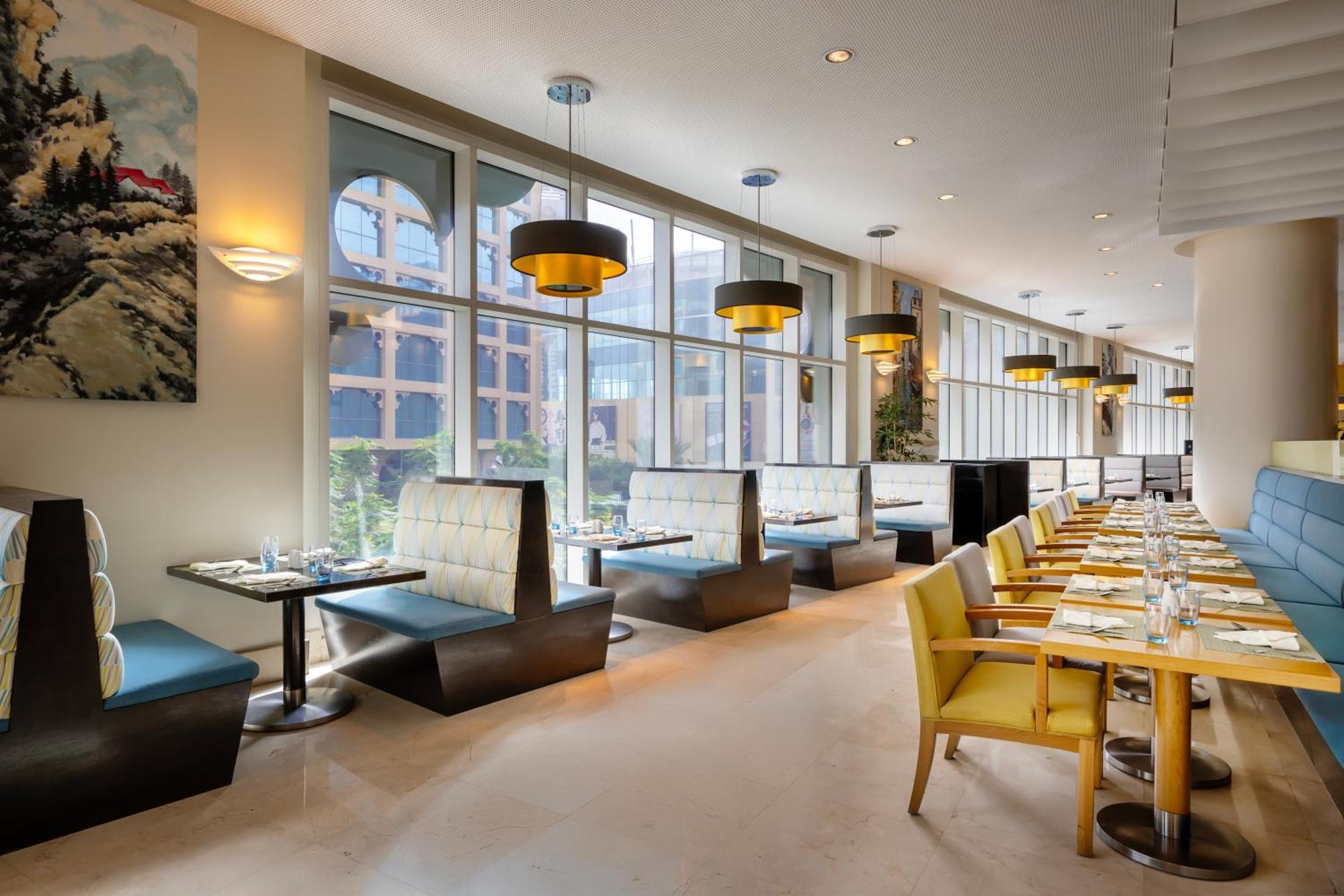 Grand Millennium Al Wahda Hotel And Executive Apartments Abu Dhabi Exterior photo The photo depicts an interior of a modern restaurant. The space features a combination of seating arrangements, including booths with cushioned backs and tables, as well as some yellow chairs. Large windows allow plenty of natural light to flood the 