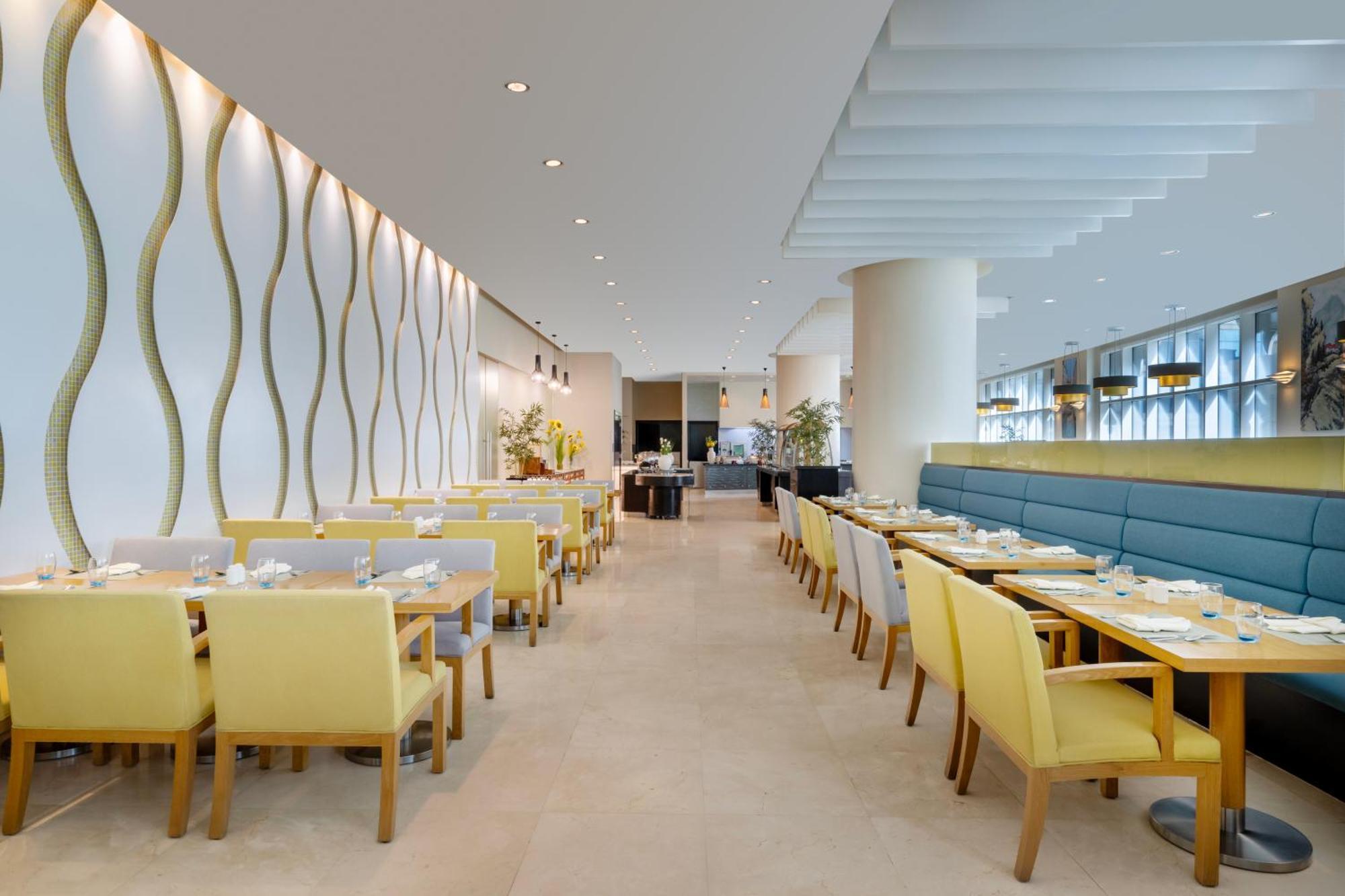 Grand Millennium Al Wahda Hotel And Executive Apartments Abu Dhabi Exterior photo The photo shows a modern restaurant interior. There are tables arranged in rows with yellow wooden chairs. The walls feature a stylish design with wavy patterns, and there are large windows allowing natural light to brighten the space. Some greenery 