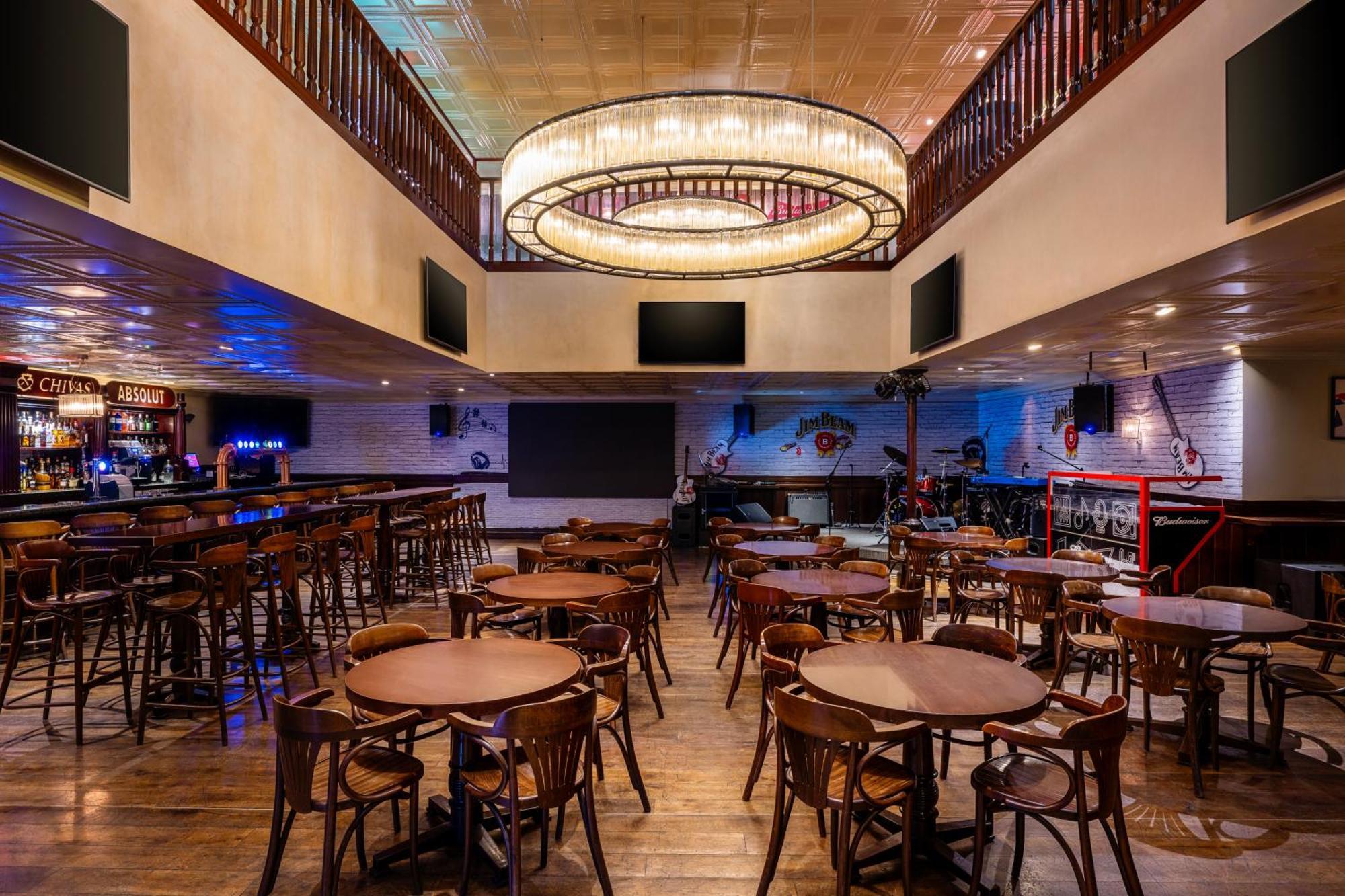 Grand Millennium Al Wahda Hotel And Executive Apartments Abu Dhabi Exterior photo The photo depicts the interior of a bar or restaurant. It features a spacious layout with several round wooden tables and chairs arranged throughout the area. There is a prominent chandelier hanging from the ceiling, providing ambient lighting. The e