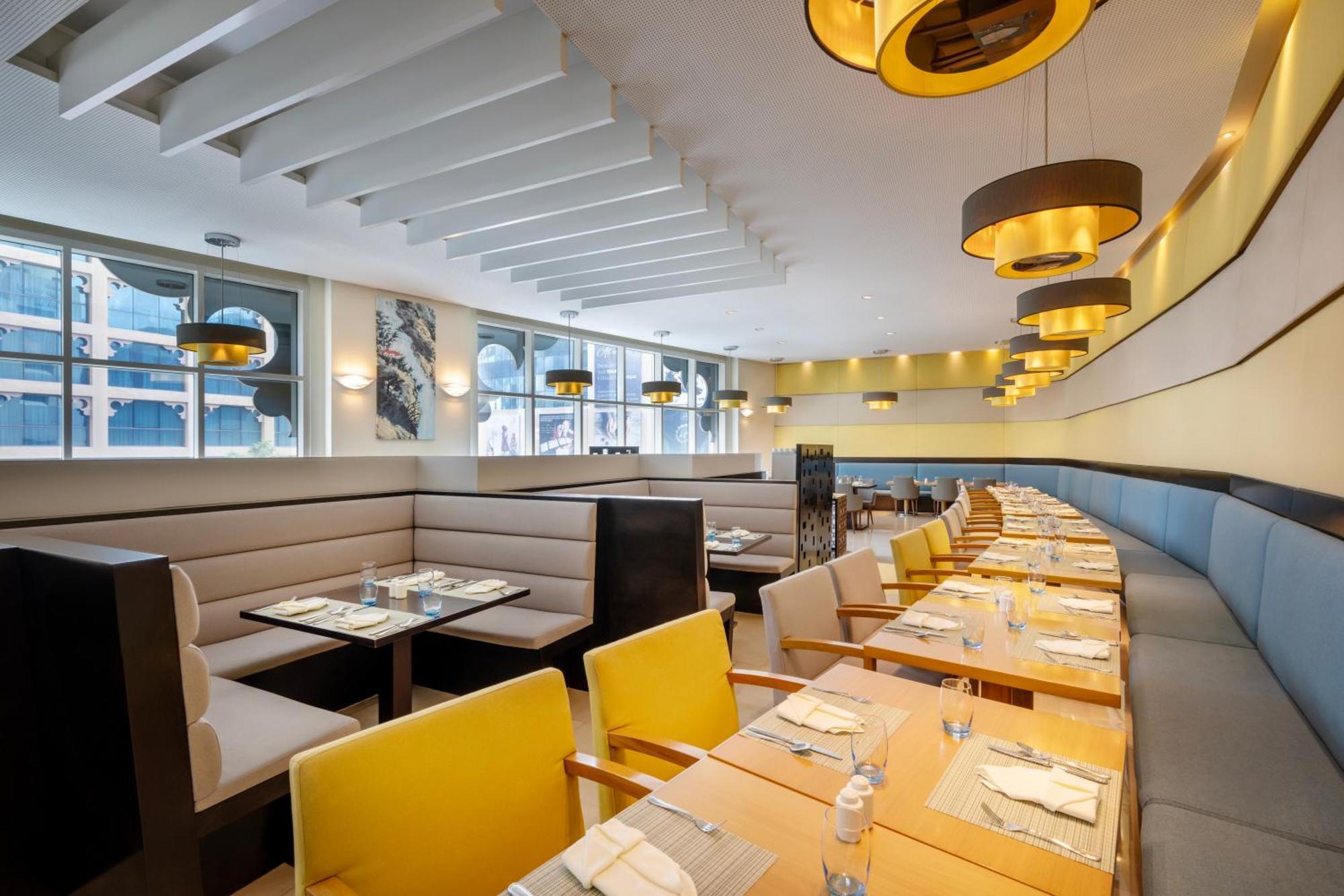 Grand Millennium Al Wahda Hotel And Executive Apartments Abu Dhabi Exterior photo The photo shows the interior of a modern restaurant. It features a clean and contemporary design with a bright atmosphere. There are multiple tables set up with yellow chairs and wooden surfaces, arranged in a long line. The seating includes a combin