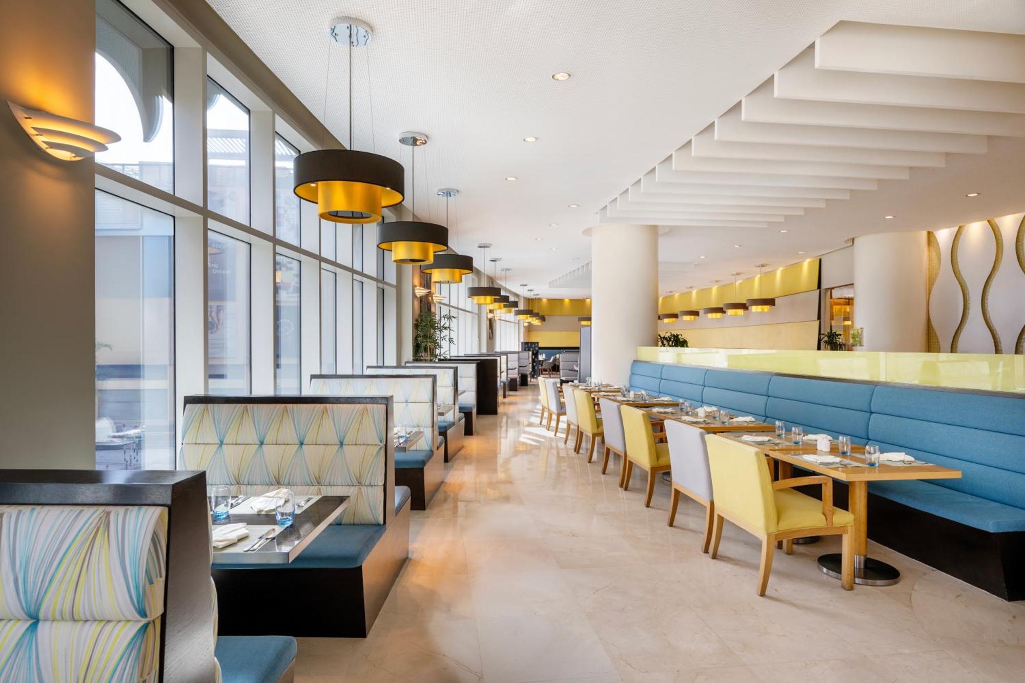 Grand Millennium Al Wahda Hotel And Executive Apartments Abu Dhabi Exterior photo The photo depicts the interior of a modern restaurant. It features large windows that allow natural light to flood the space, creating a bright and inviting atmosphere. The seating consists of elegant booths with blue and gray upholstery, as well as 