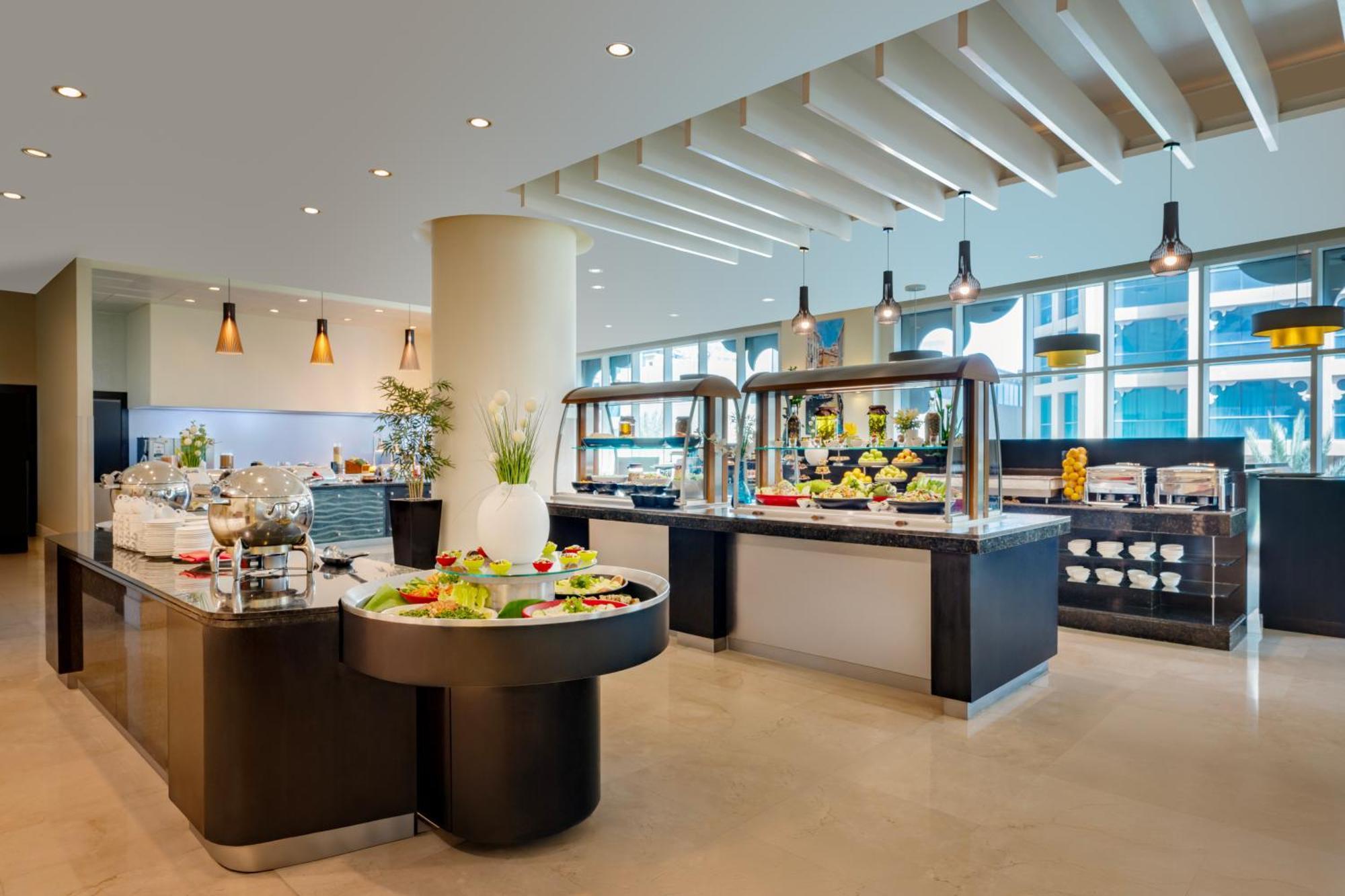 Grand Millennium Al Wahda Hotel And Executive Apartments Abu Dhabi Exterior photo The image shows a modern dining area, likely in a hotel or restaurant, featuring a buffet setup. There are several food stations with elegantly arranged dishes, including fruits and salads. The space is well-lit with large windows allowing natural li